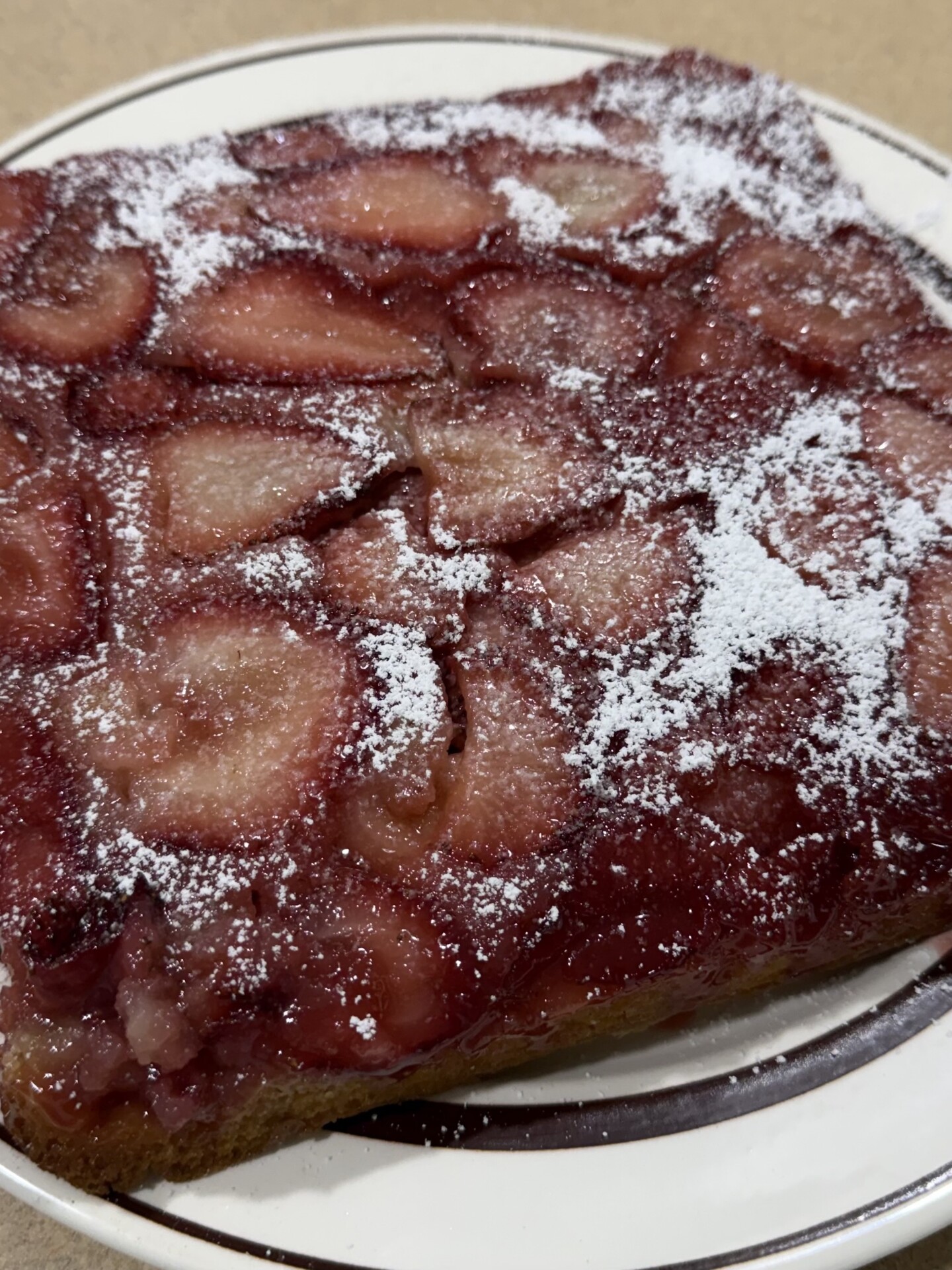 A close-up of a square strawberry upside-down cake topped with powdered sugar, served on a white plate with a brown stripe around the edge.