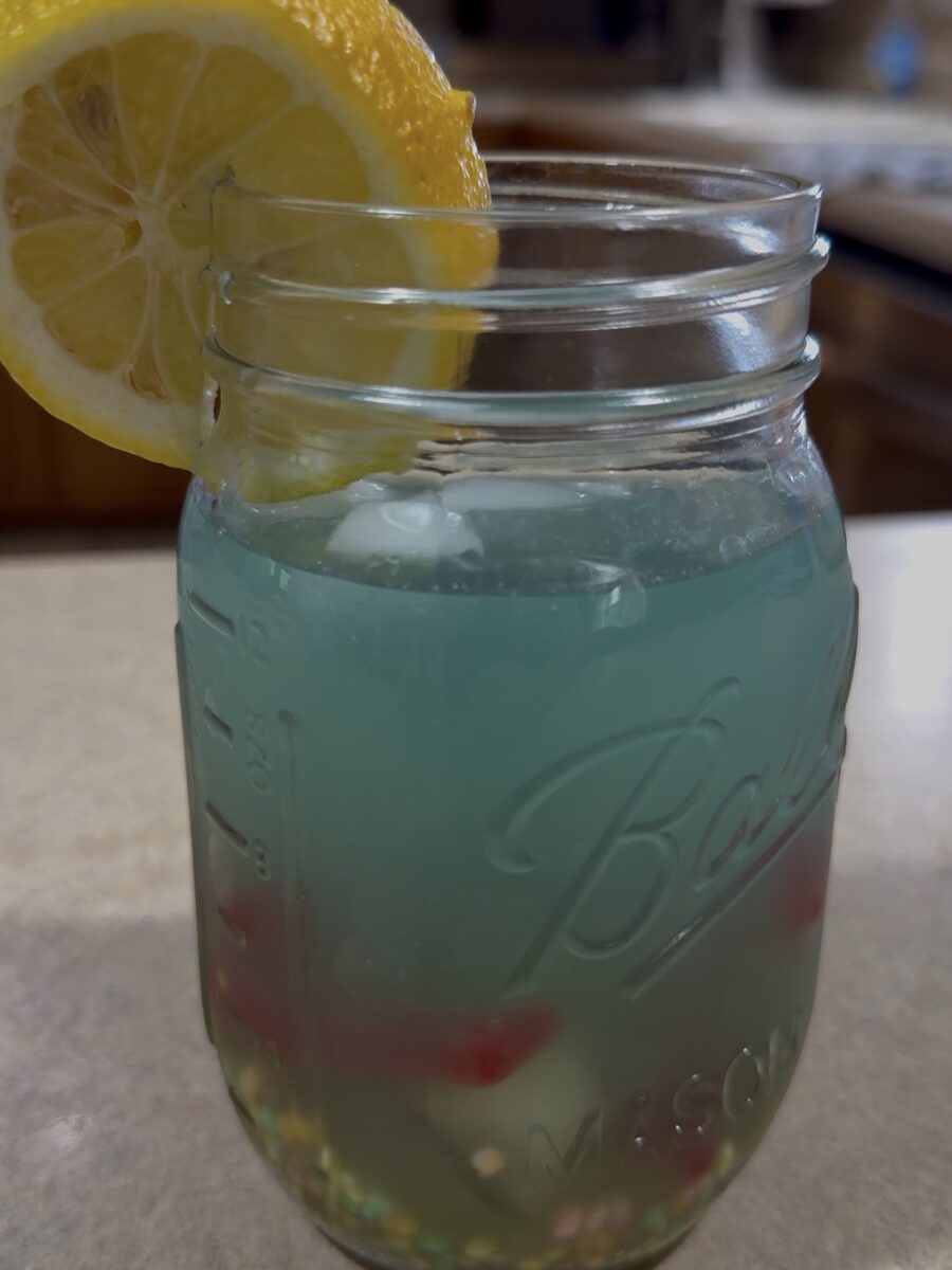 A mason jar filled with a blue-green beverage, ice, and colorful fruit pieces, garnished with a lemon slice on the rim. The jar sits on a kitchen counter.