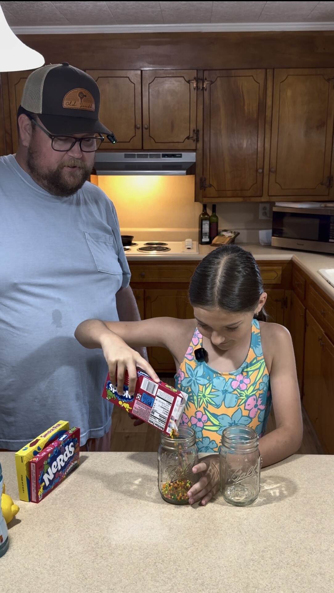 A man in a cap and glasses watches as a young girl in a floral swimsuit pours Nerds candy from a box into a glass jar on a kitchen counter. Two more empty jars and another candy box are nearby.