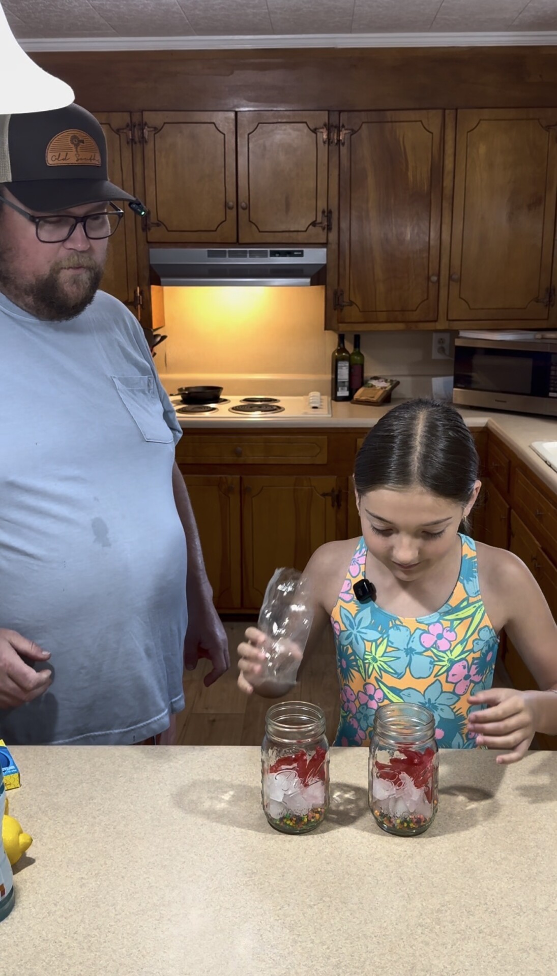 A man in a blue shirt and cap stands in a kitchen watching a young girl in a floral swimsuit pour something into a jar filled with red fruit and ice on the counter.