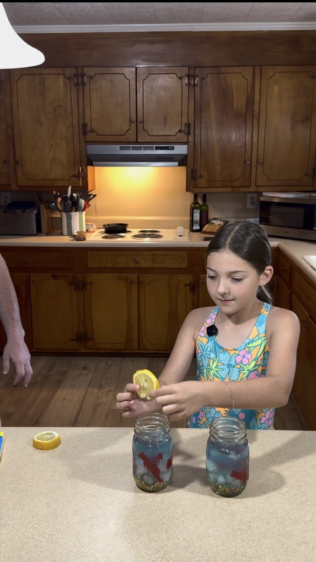 A girl in a colorful dress places a lemon slice into a mason jar filled with a blue drink and gummy candies, standing in a kitchen with wooden cabinets.