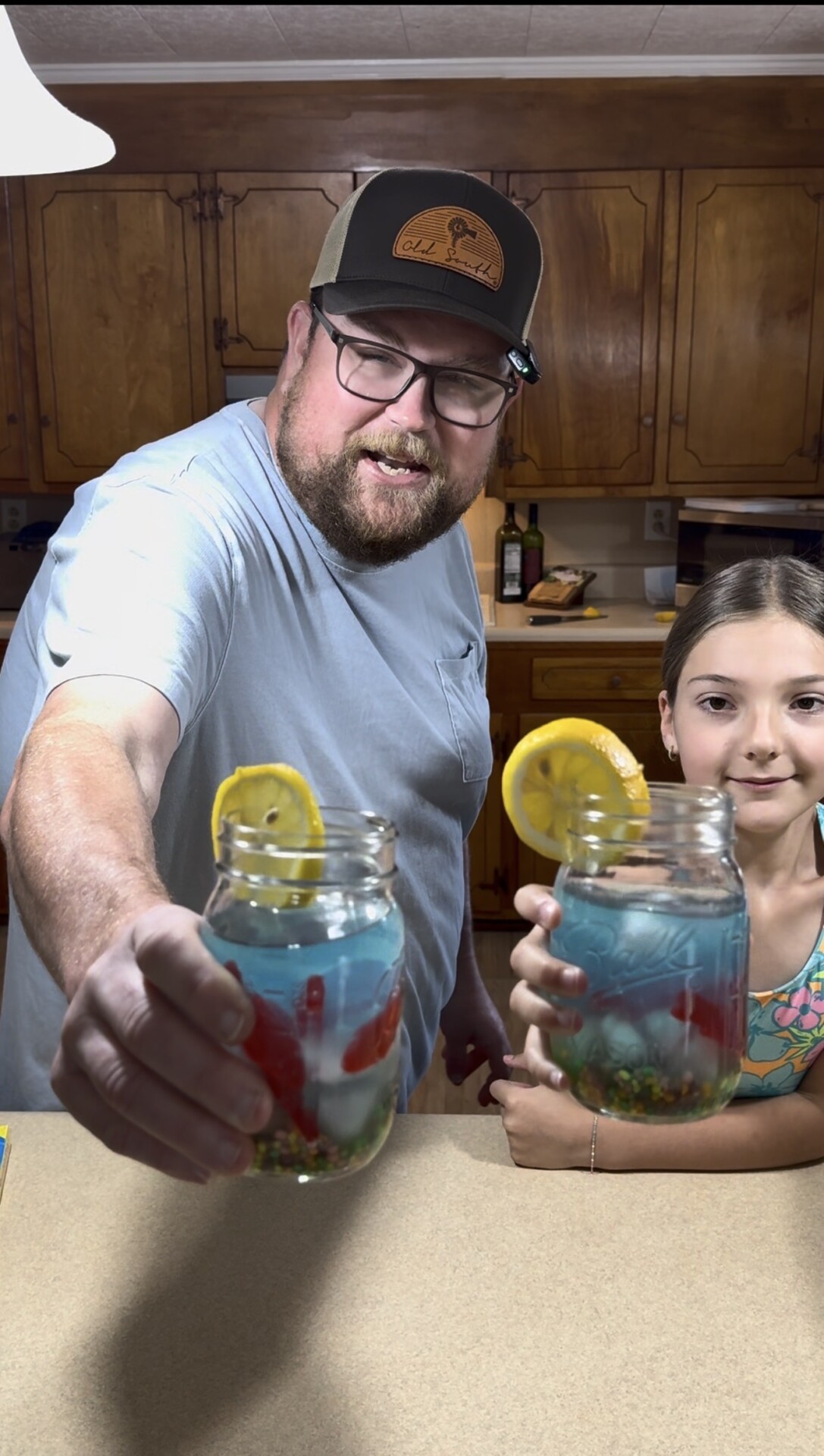 A bearded man and a young girl smile while holding up mason jars filled with a blue drink, ice, fruit, and lemon slices, standing in a kitchen with wooden cabinets.