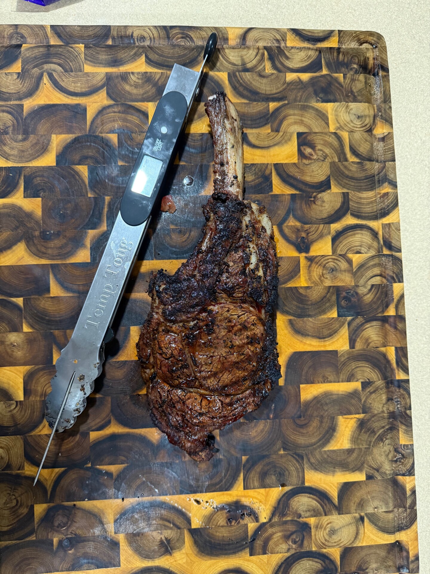 A cooked tomahawk steak rests on a patterned wooden cutting board next to a metal meat thermometer displaying a digital reading.