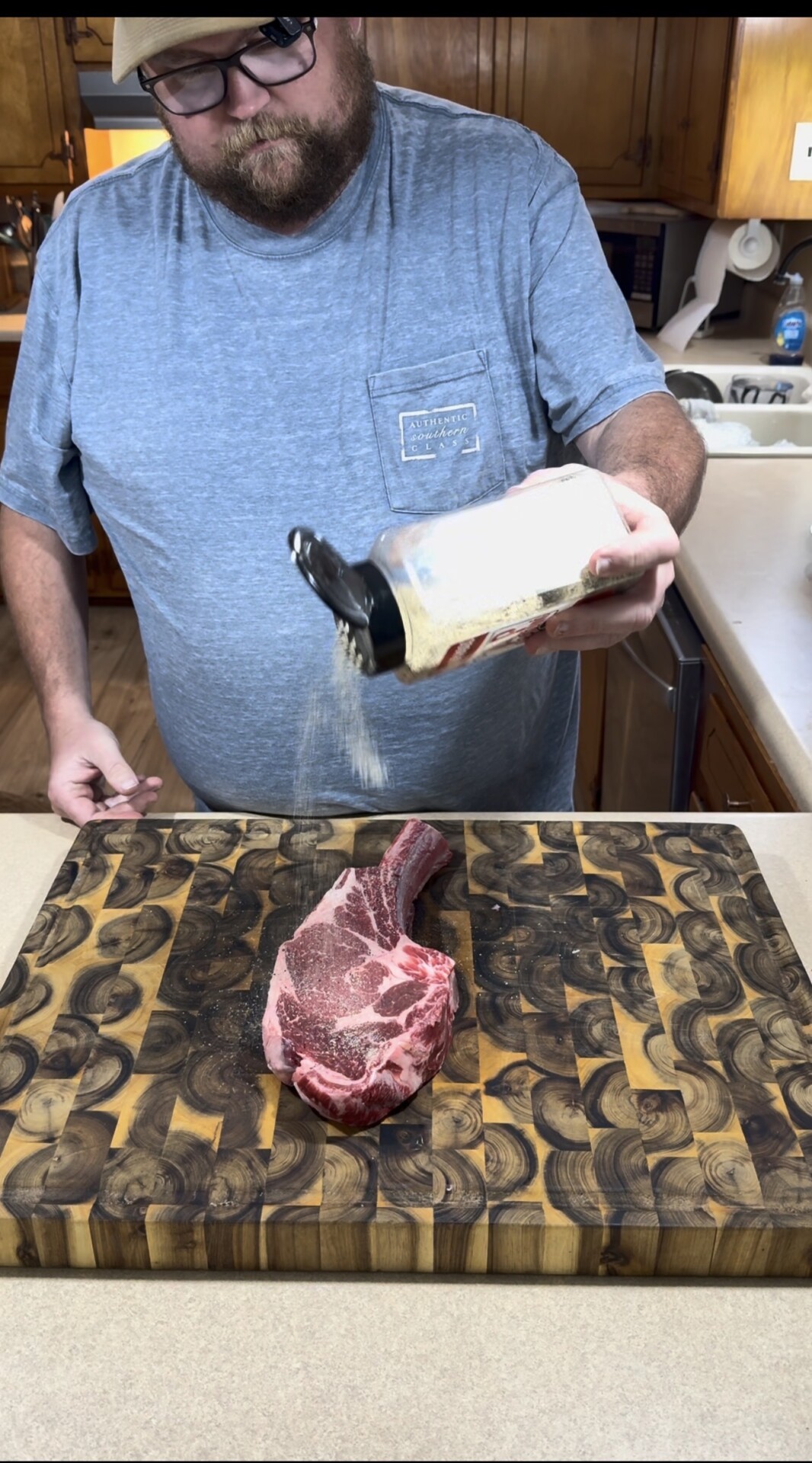A person in a blue t-shirt is seasoning a raw tomahawk steak on a patterned wooden cutting board in a kitchen, pouring seasoning from a large container.