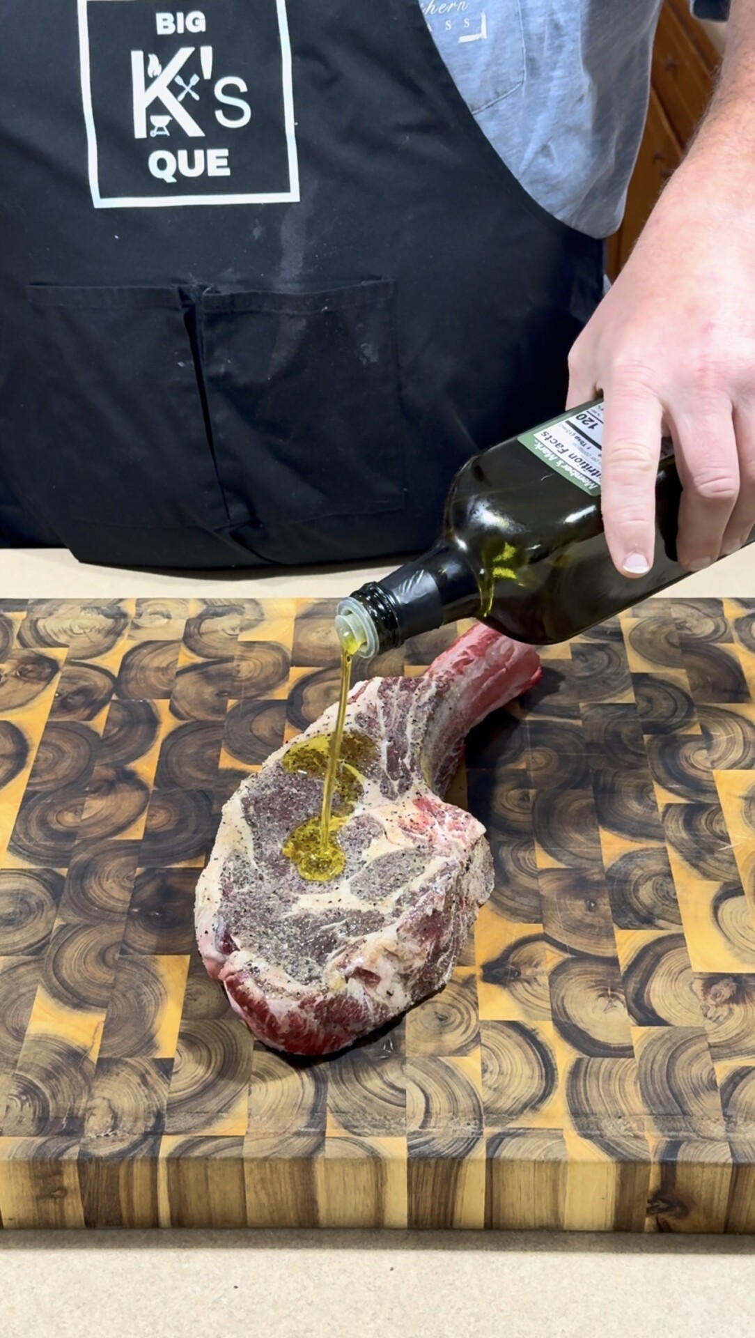 A person wearing a black apron pours olive oil onto a seasoned tomahawk steak resting on a wooden cutting board.