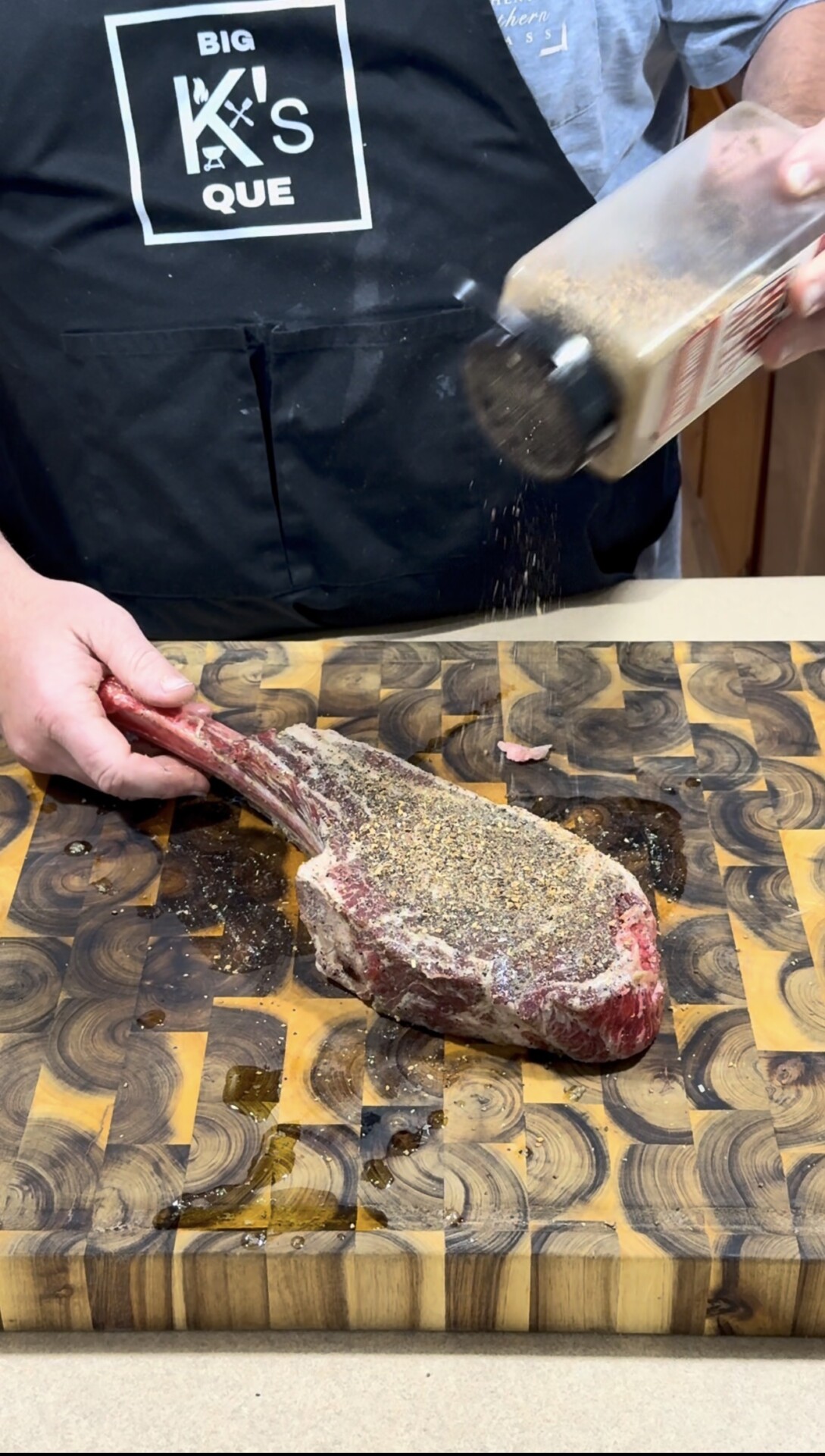 A person in a black apron sprinkles seasoning onto a large raw tomahawk steak on a patterned wooden cutting board.