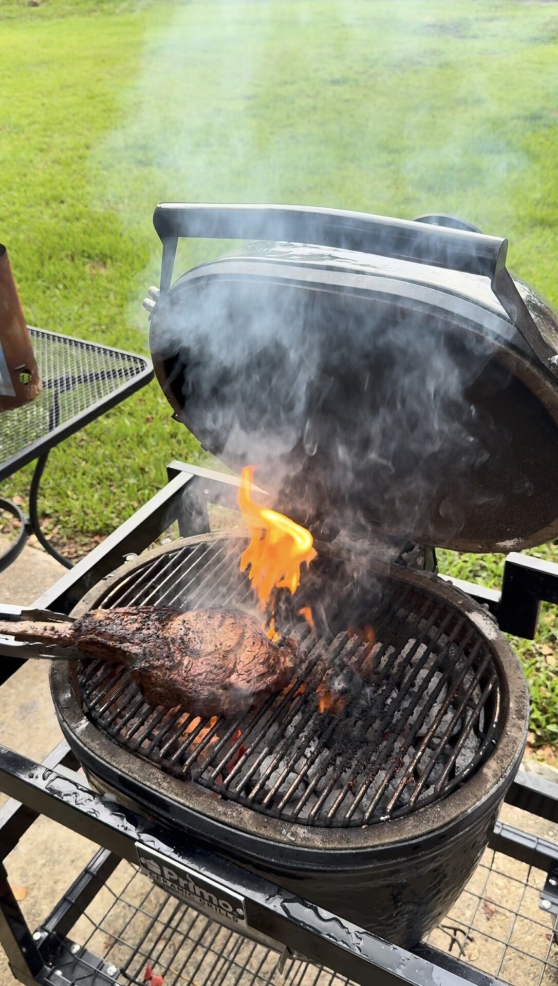 A tomahawk steak is cooking on a charcoal grill with the lid open, smoke rising, and a small flame touching the steak. The grill is set outdoors on a patio beside a green lawn.