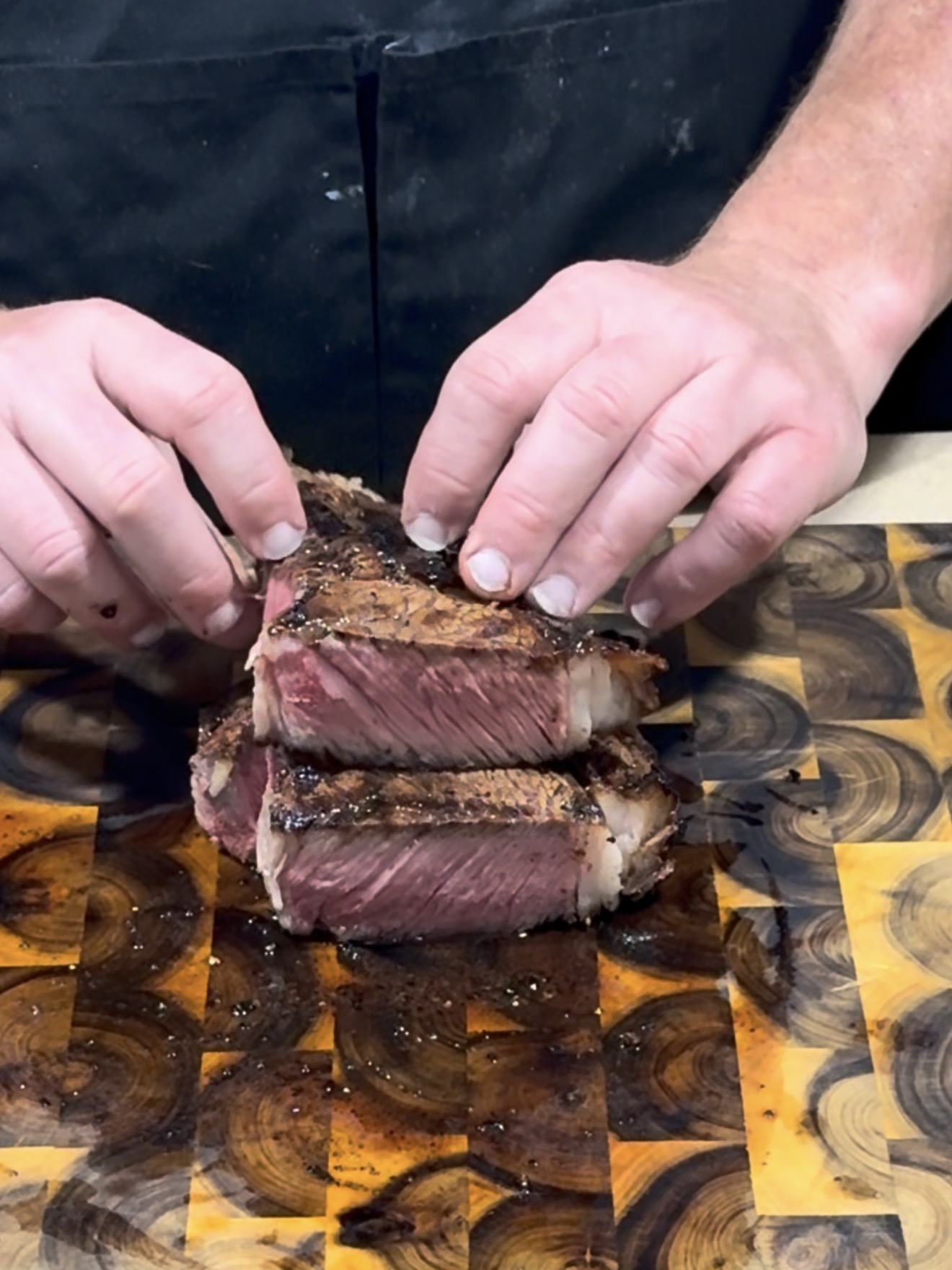 A person places two thick, juicy slices of grilled steak on top of each other on a wooden cutting board. The steak is cooked medium rare, with a smoked outer crust and a pink interior.