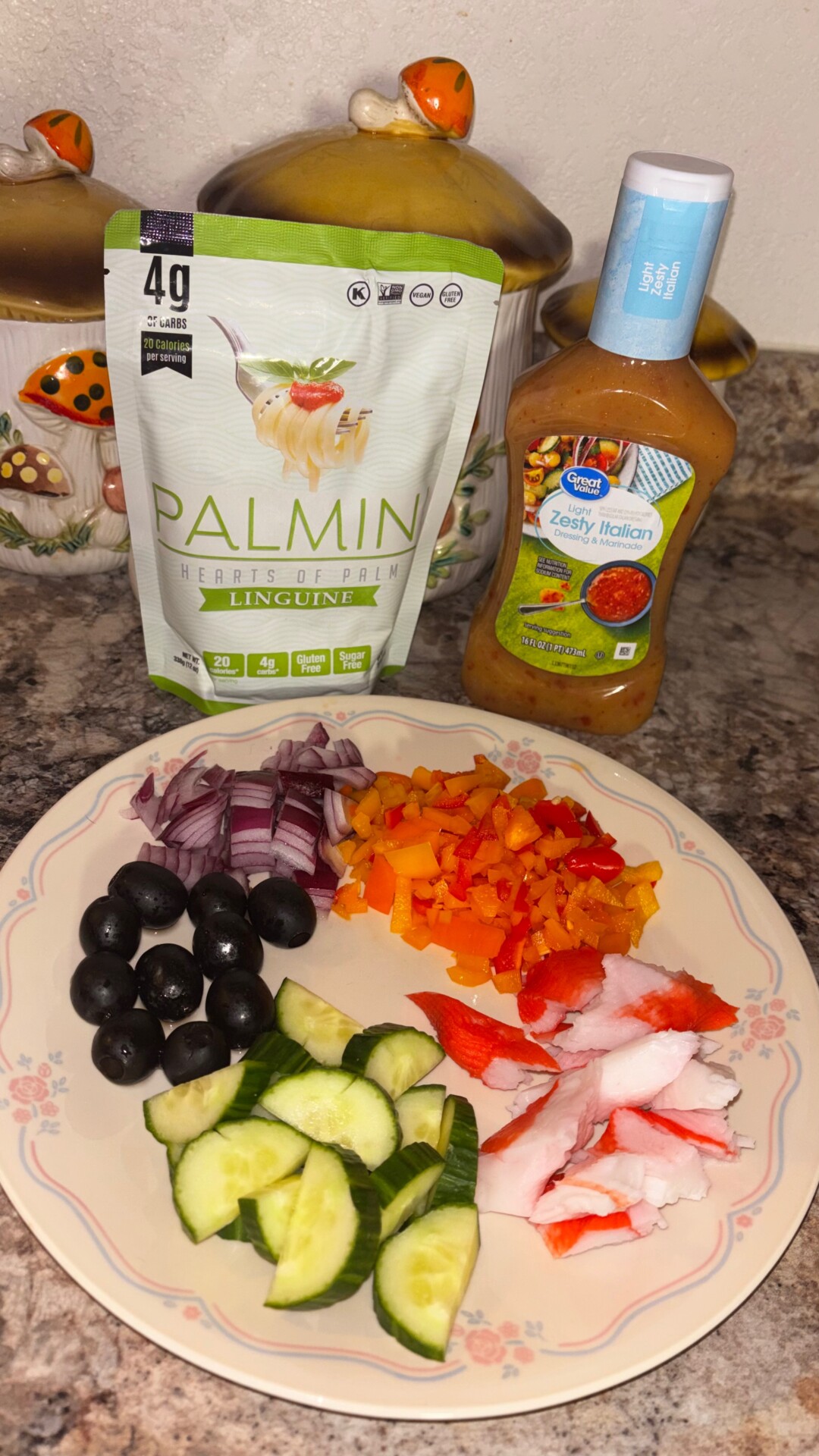 A plate with sliced cucumbers, black olives, crab sticks, chopped red onion, and diced red and orange bell peppers sits on a counter next to a bag of Palmini linguine and a bottle of zesty Italian dressing.