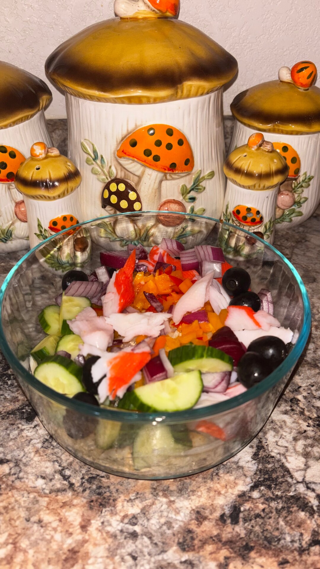 A glass bowl filled with salad ingredients including cucumbers, olives, onion, and imitation crab sits on a granite countertop in front of mushroom-themed ceramic canisters.