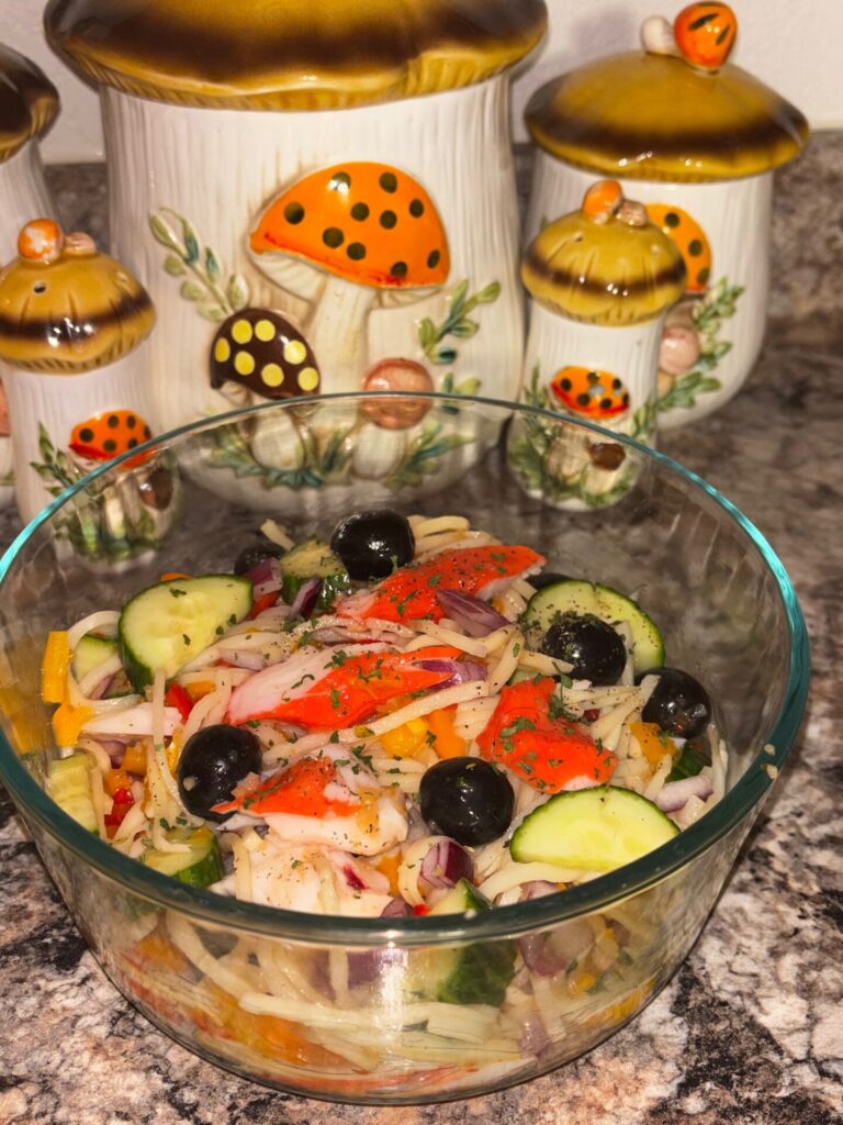 A glass bowl of colorful salad with imitation crab, cucumbers, olives, onions, and shredded cheese sits on a kitchen counter in front of three ceramic mushroom-shaped canisters.
