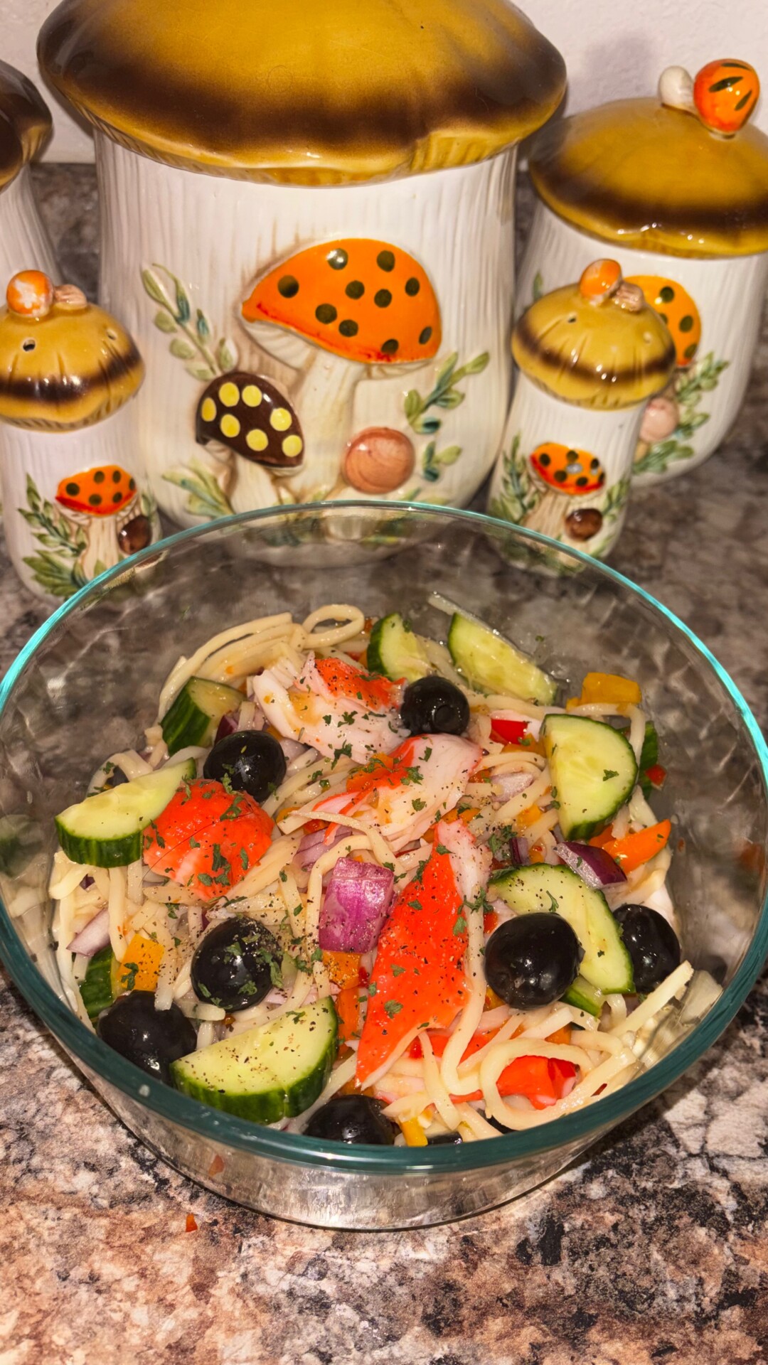 A glass bowl of pasta salad with cucumber, red onion, olives, bell pepper, and imitation crab sits on a granite countertop in front of ceramic kitchen canisters decorated with mushrooms.
