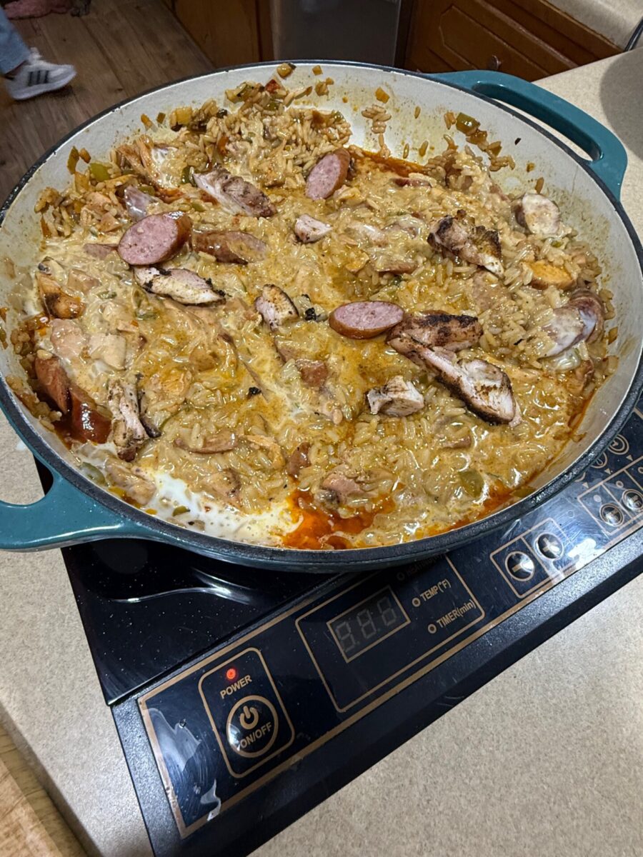 A large round pan on a stovetop contains a creamy, saucy dish with sliced sausage, chicken, onions, and rice. The food looks partially stirred, with some browning and melted cheese on top.