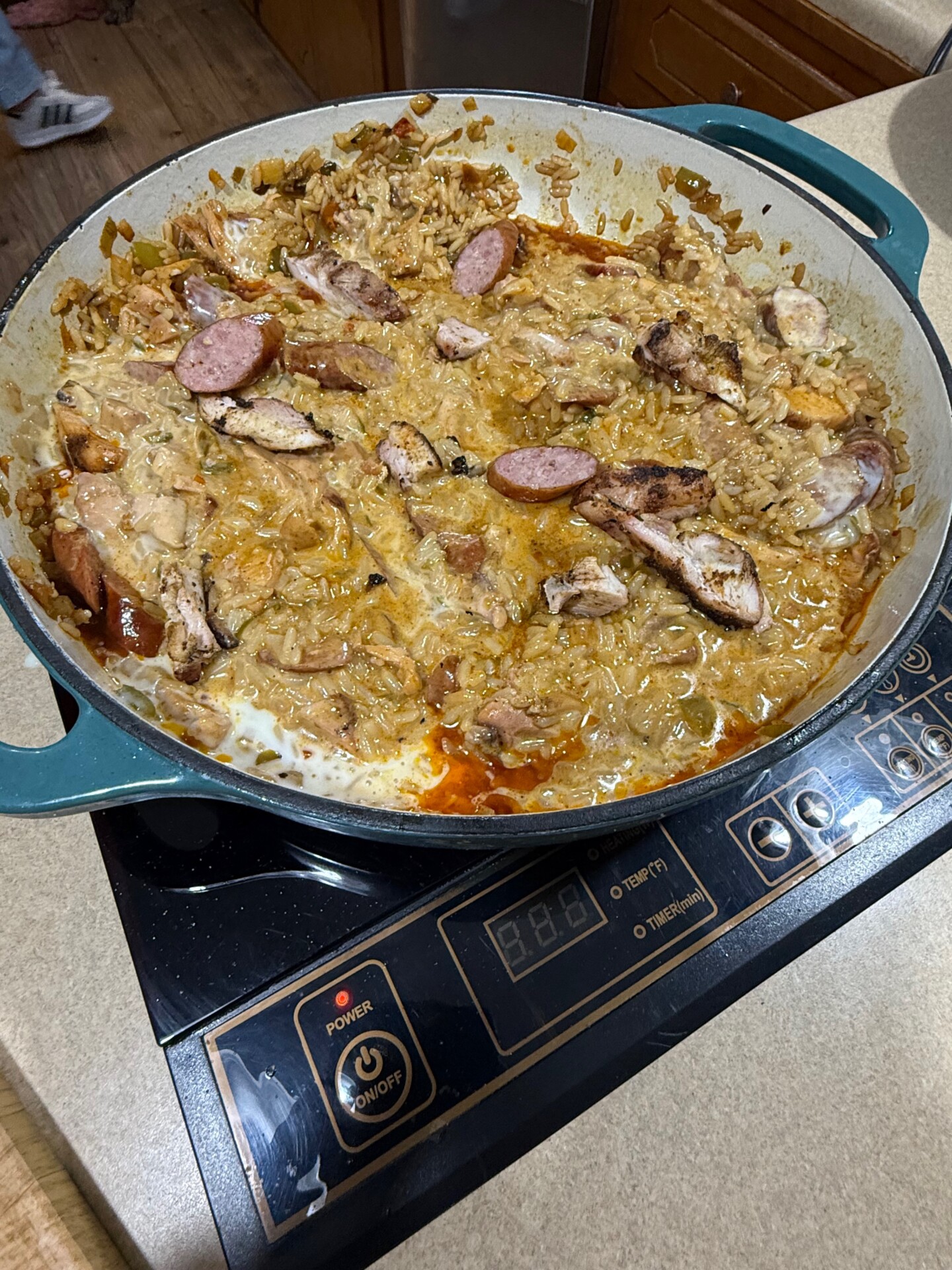 A large round pan on a stovetop contains a creamy, saucy dish with sliced sausage, chicken, onions, and rice. The food looks partially stirred, with some browning and melted cheese on top.