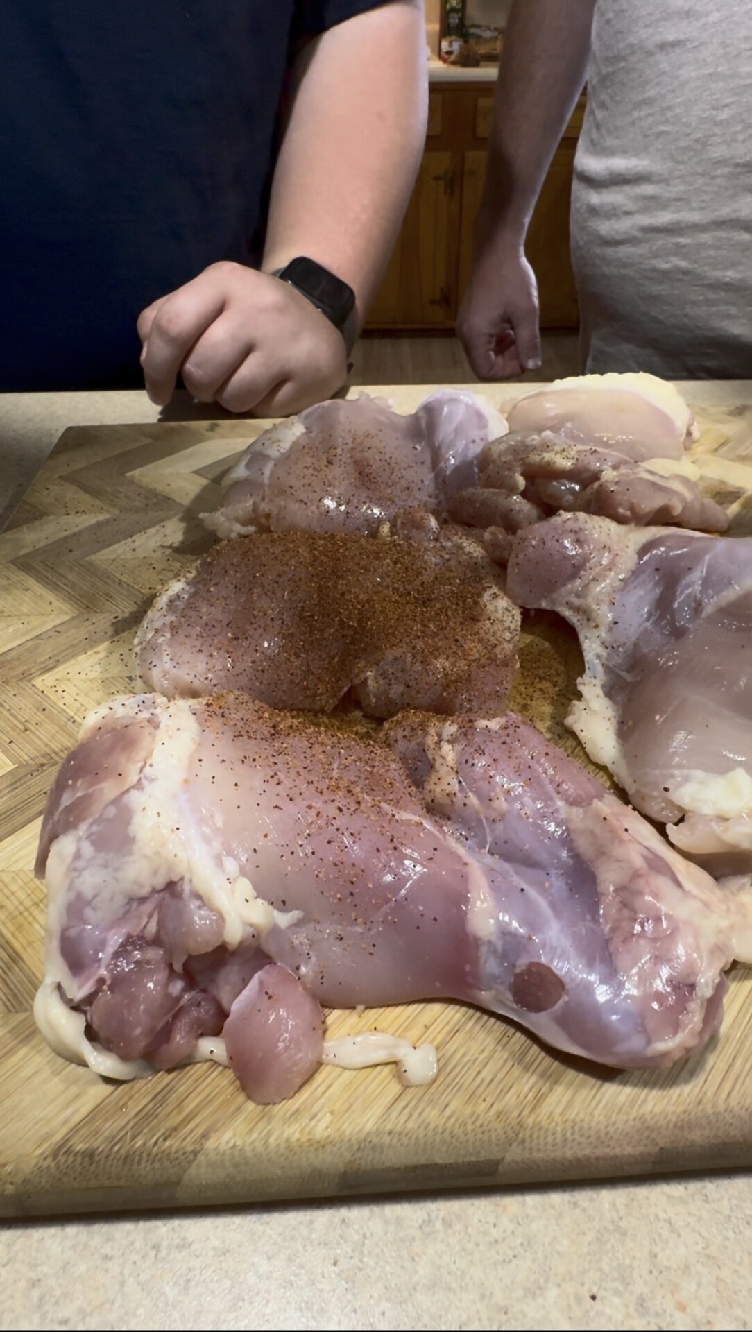 Two people stand behind a wooden cutting board covered with raw chicken pieces being seasoned with a brown spice blend. One person wears a watch and dark shirt; the other is in a light shirt.
