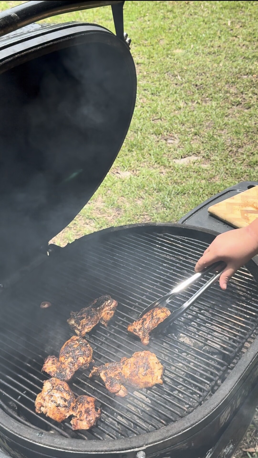 A person uses tongs to grill seasoned chicken pieces on a smoky charcoal barbecue grill outdoors, with green grass visible in the background.