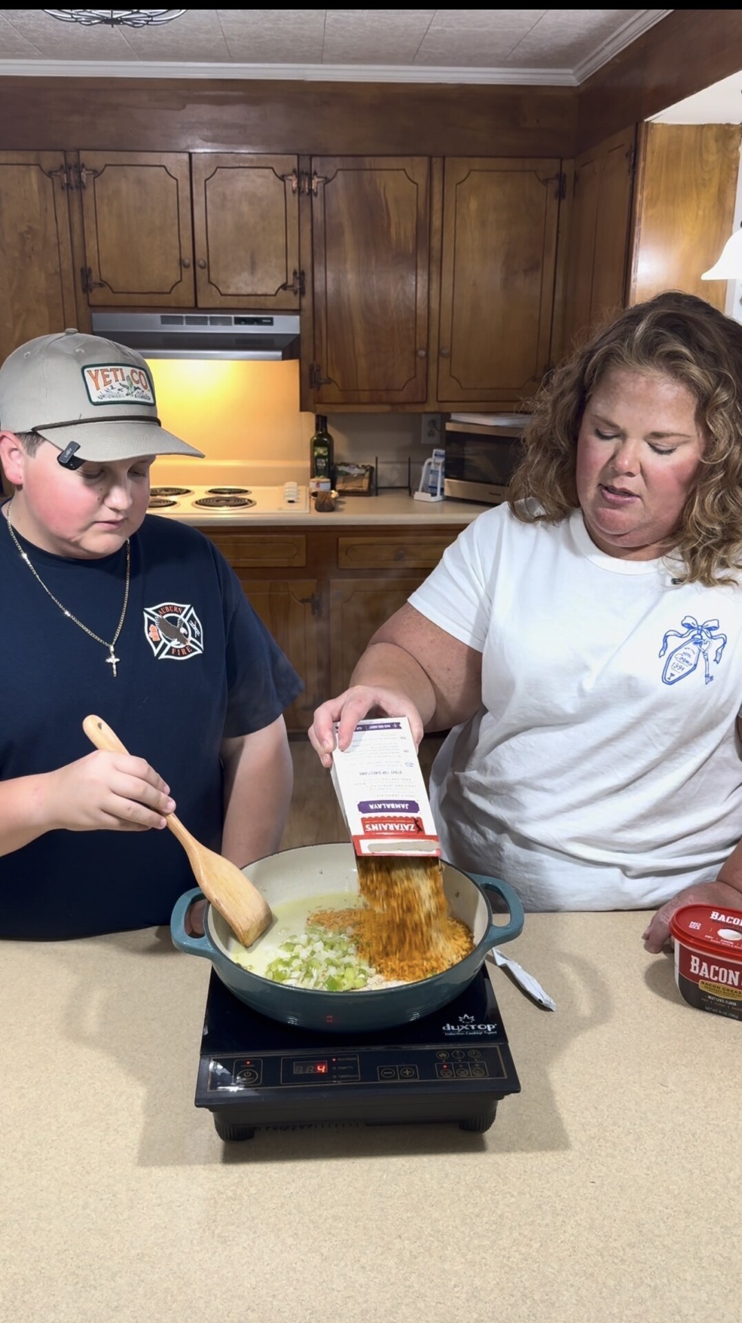 A woman pours a box of pasta into a pot on an electric stove while a boy stirs the contents. They are cooking together in a kitchen with wooden cabinets. A container of bacon bits sits on the counter.