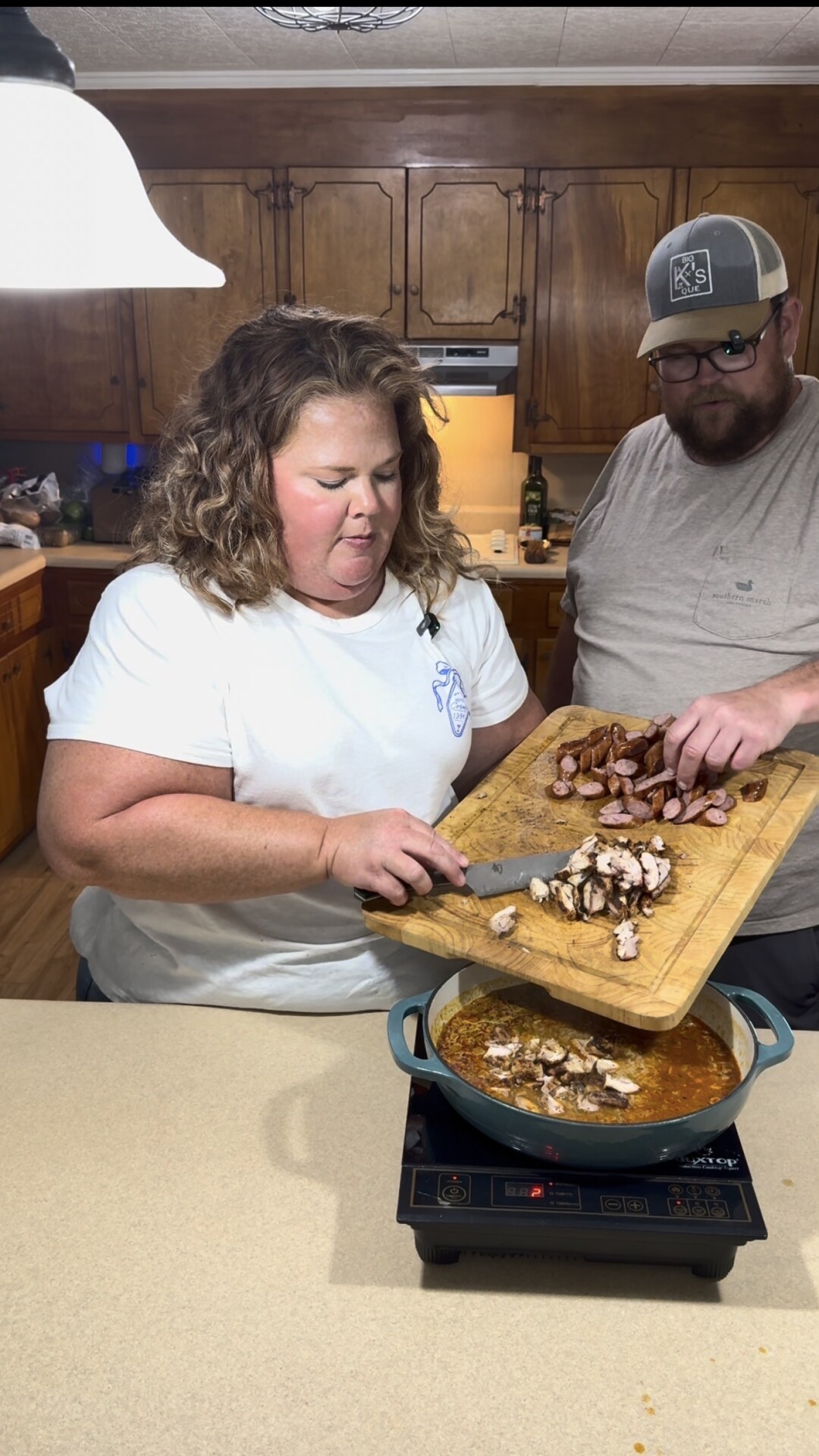 A woman in a white shirt adds sliced sausage and chicken from a cutting board into a pot of stew on a countertop, while a man in a cap and glasses stands beside her in a kitchen.