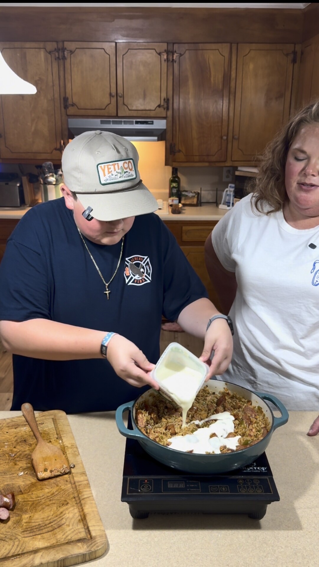 A boy in a cap pours a creamy white sauce into a pan of food on a kitchen counter while a woman in a white shirt stands beside him, watching.
