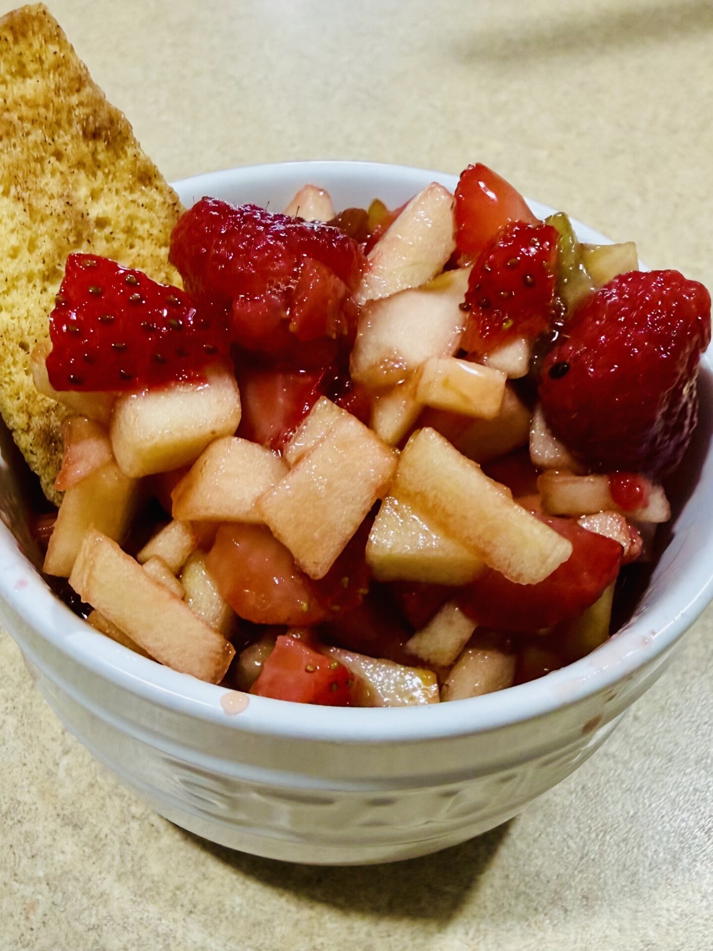 A white bowl filled with a fruit salad made of chopped apples, strawberries, and other fruits, garnished with a biscuit on the side, sitting on a beige countertop.