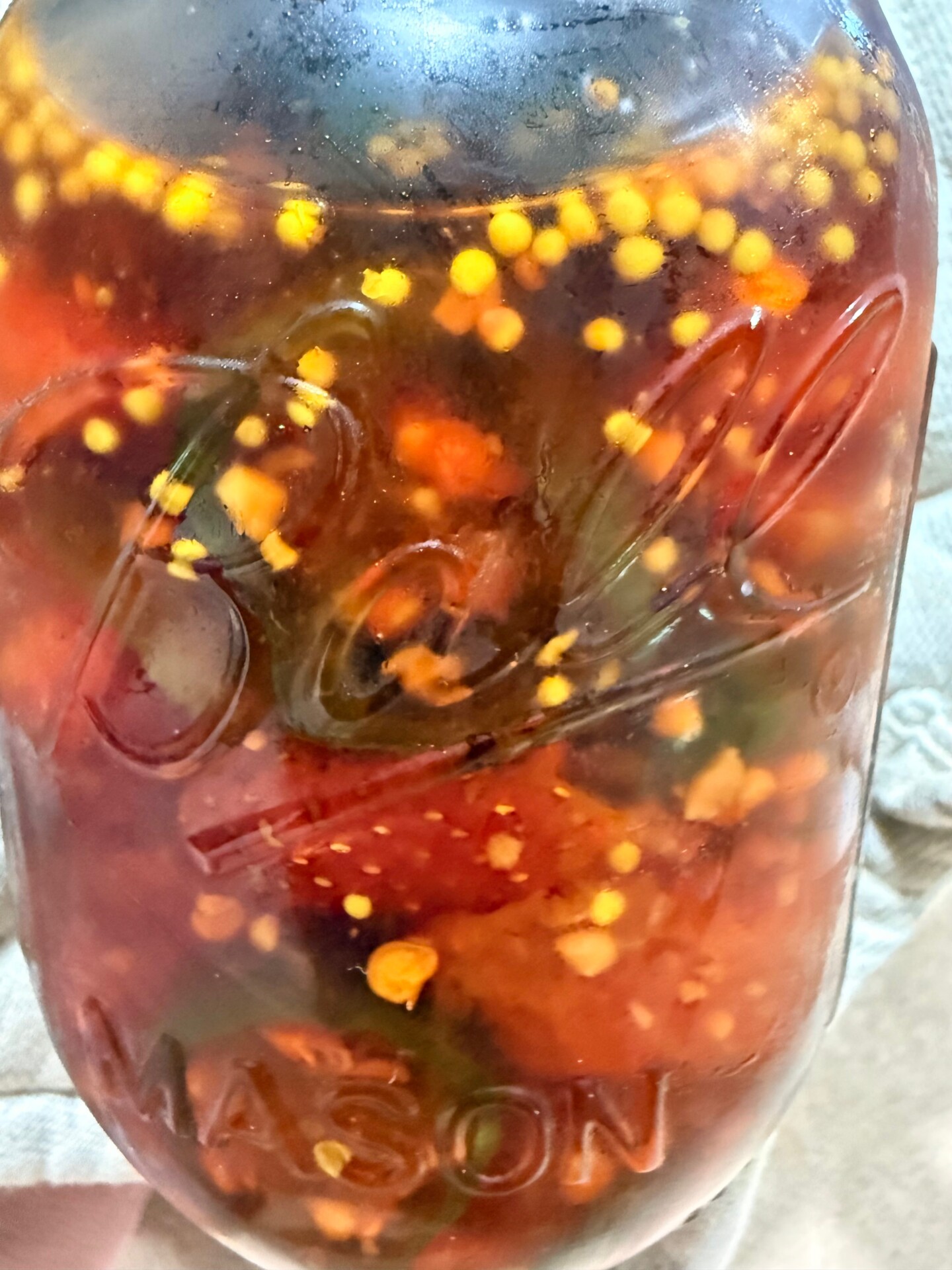 A close-up of a Ball Mason jar filled with colorful pickled vegetables and floating yellow spices in a brine. The jar is held up, and the contents are visible through the glass.