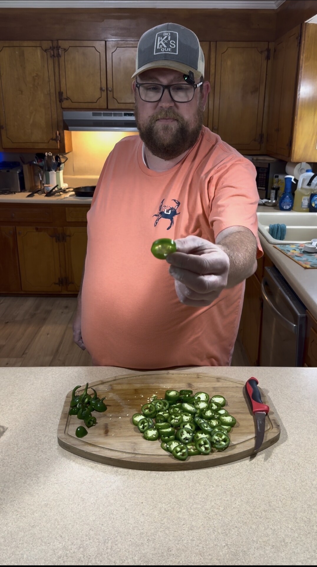 A man in a peach shirt and cap stands in a kitchen, holding a sliced jalapeño pepper toward the camera. A cutting board with more sliced jalapeños and a knife sits on the counter in front of him.