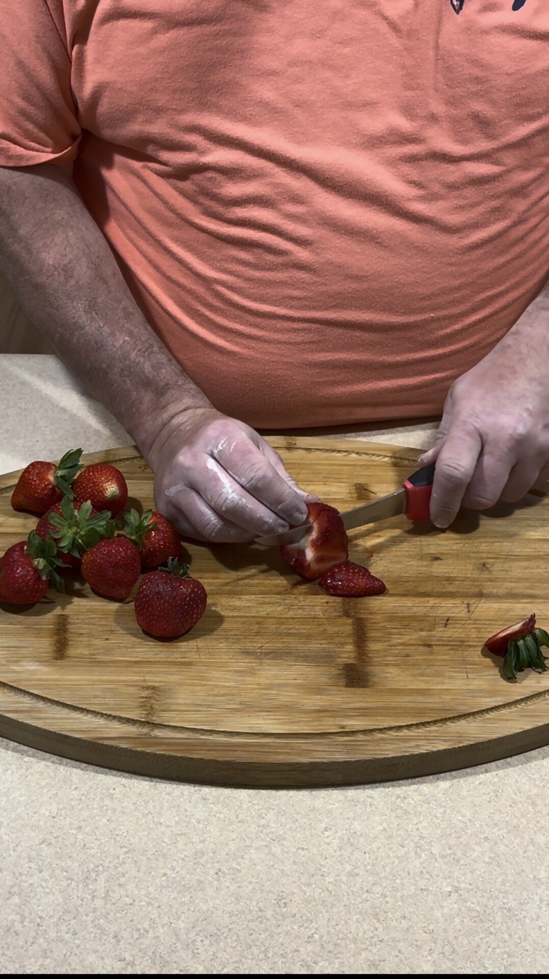 A person wearing a peach shirt slices fresh strawberries on a wooden cutting board. Several whole strawberries and green strawberry tops are also visible on the board.