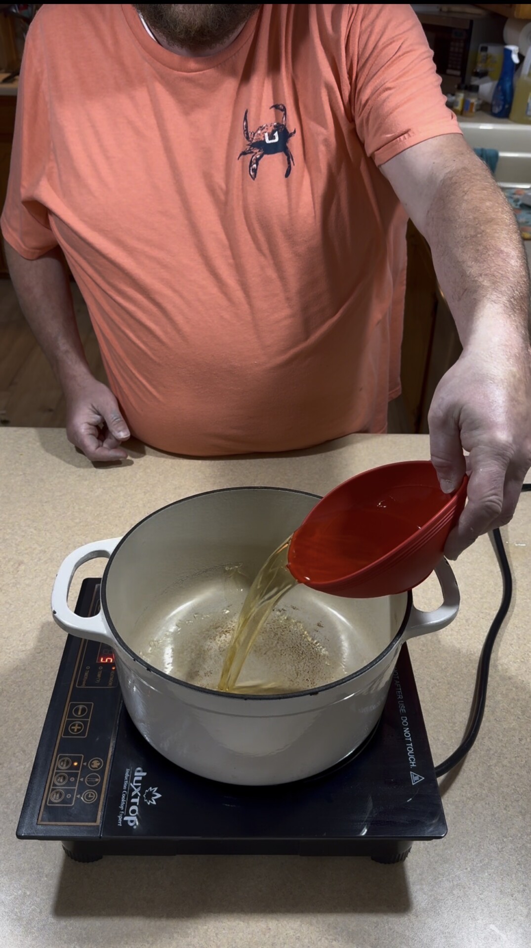 A person in an orange shirt pours liquid from a red measuring cup into a white pot on an electric cooktop in a kitchen.