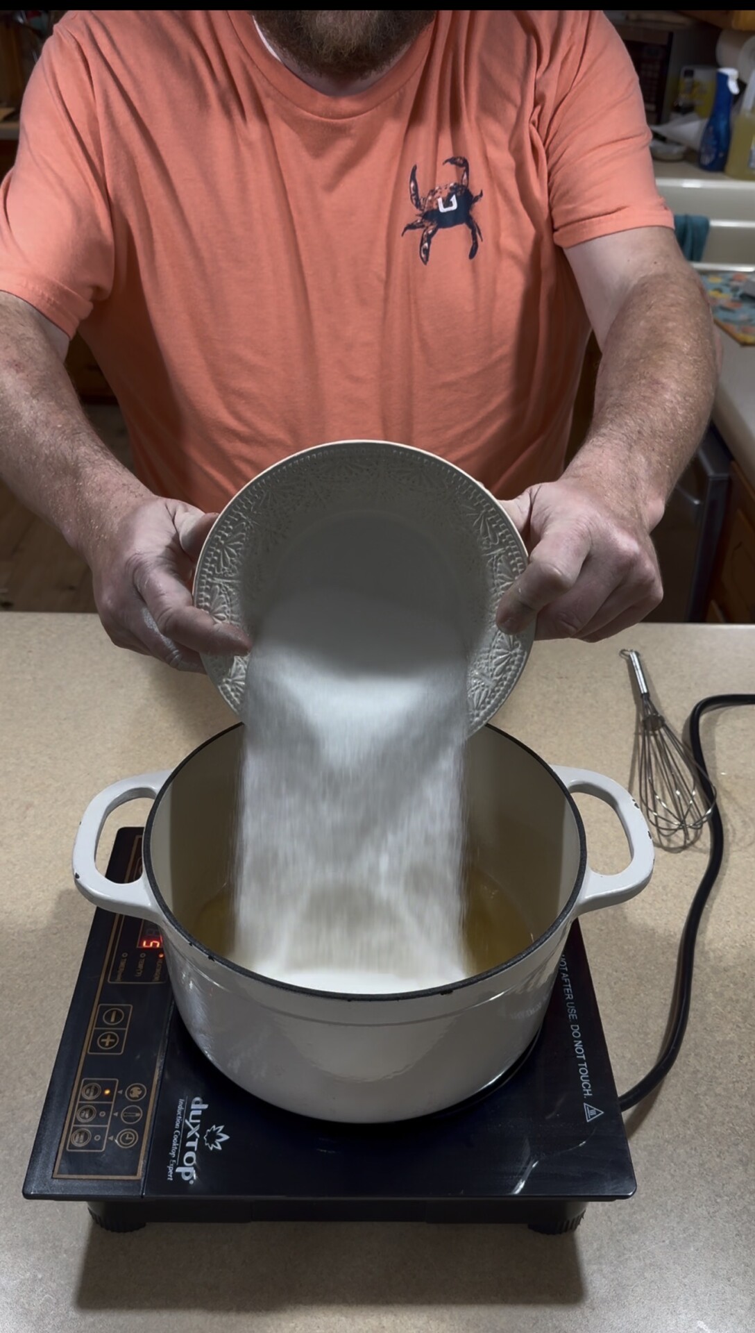 A person wearing an orange shirt pours a large amount of sugar from a white plate into a white pot on an electric stovetop. A whisk lies nearby on the kitchen counter.