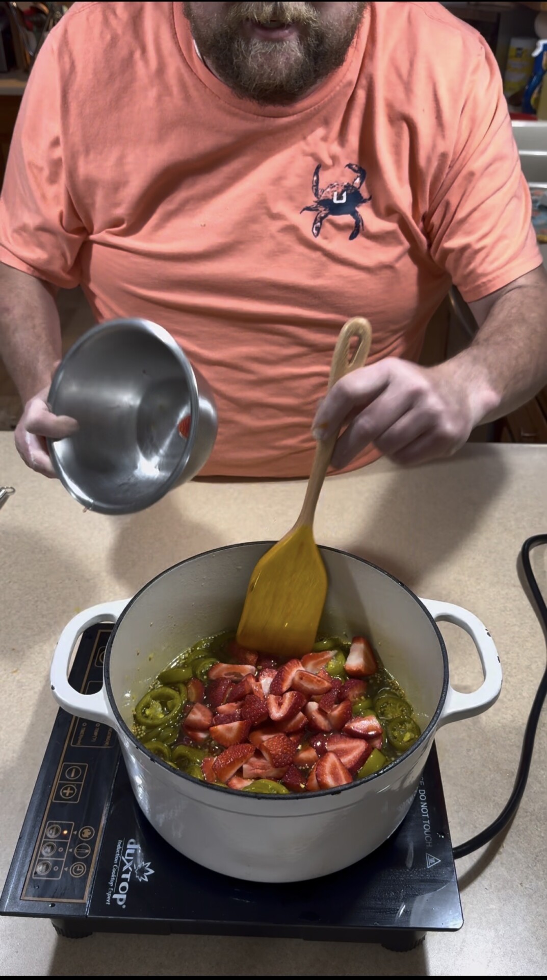 A person in an orange shirt stirs chopped strawberries and sliced green peppers in a white pot on an electric stovetop, holding a metal bowl in their other hand.