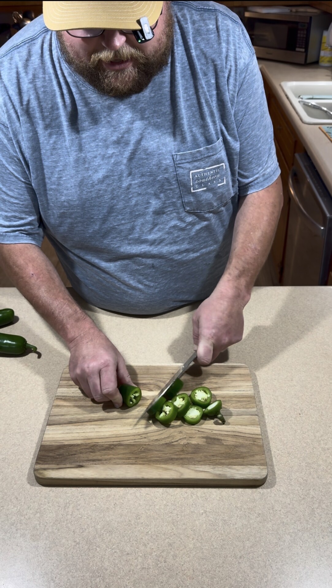 A person in a blue shirt slices fresh jalapeño peppers on a wooden cutting board on a kitchen counter. Several whole jalapeños are visible nearby.