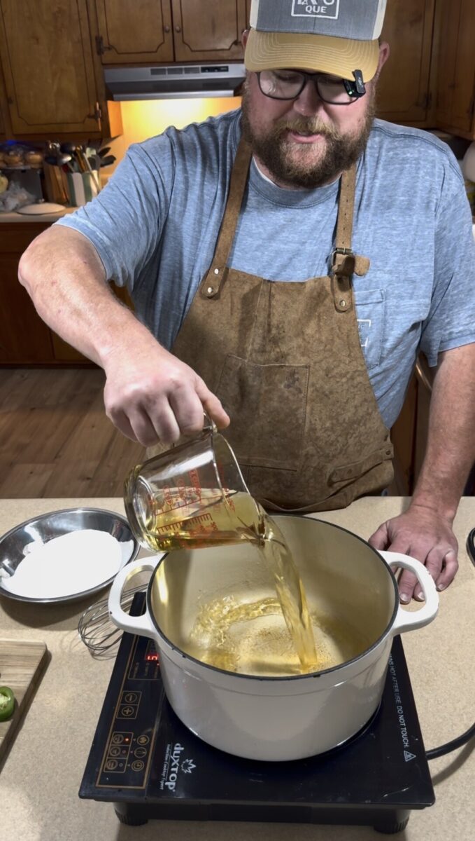 A person wearing a brown apron and blue shirt pours liquid from a measuring cup into a large white pot on an induction cooktop in a kitchen, with cooking ingredients nearby on the counter.