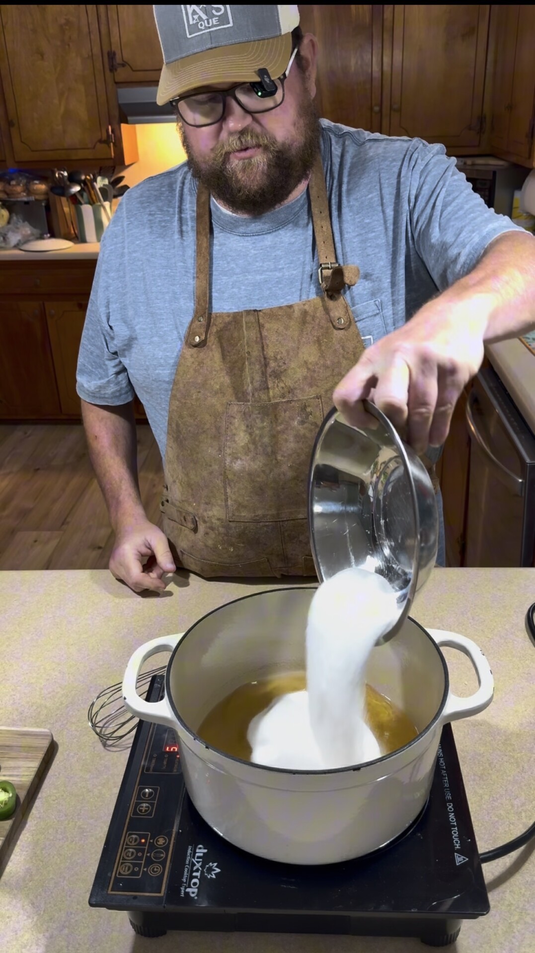 A bearded person wearing glasses, a cap, a blue shirt, and a brown apron pours a large amount of white sugar from a metal bowl into a pot on an electric stove in a kitchen.