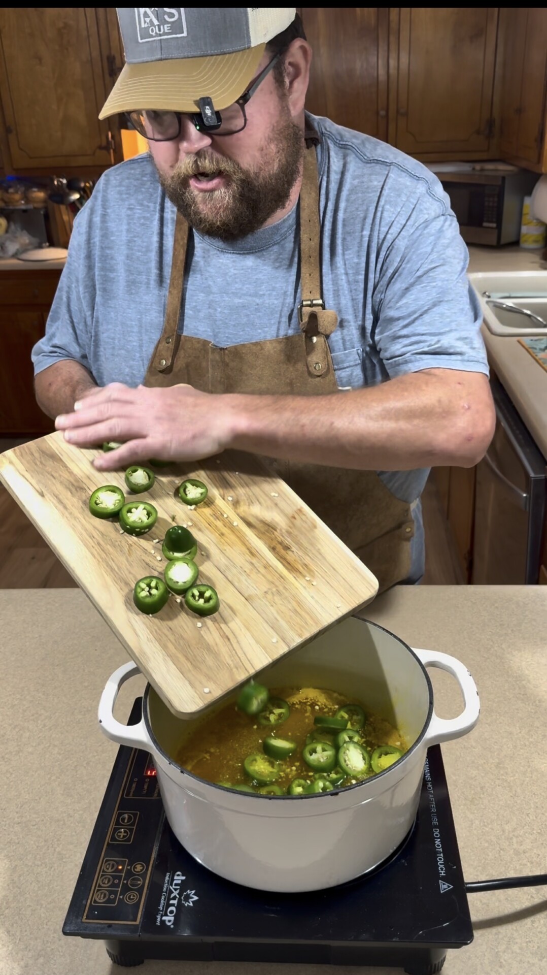A person wearing glasses, a cap, and an apron adds sliced jalapeños from a cutting board into a pot on a stovetop in a kitchen.
