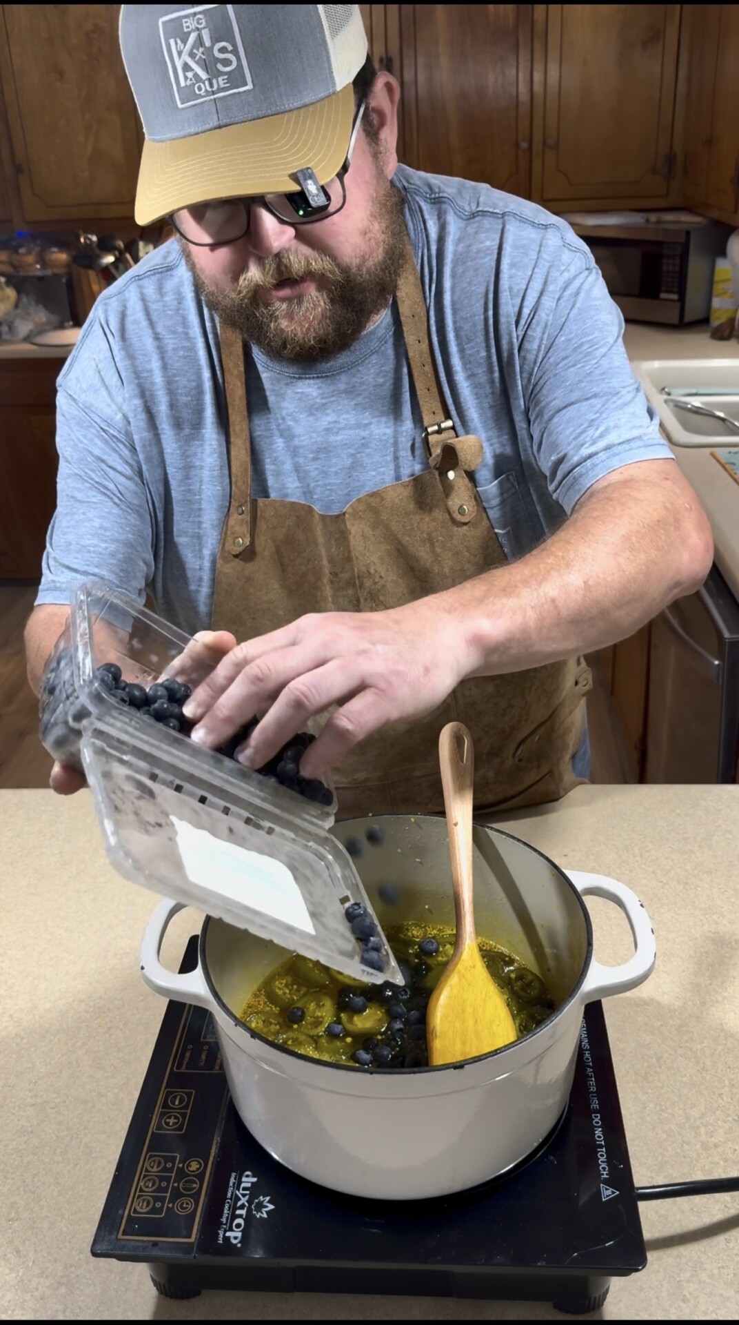 A bearded man wearing a cap, glasses, and a brown apron pours fresh blueberries from a plastic container into a white pot on a stovetop in a kitchen. A yellow spoon rests in the pot.
