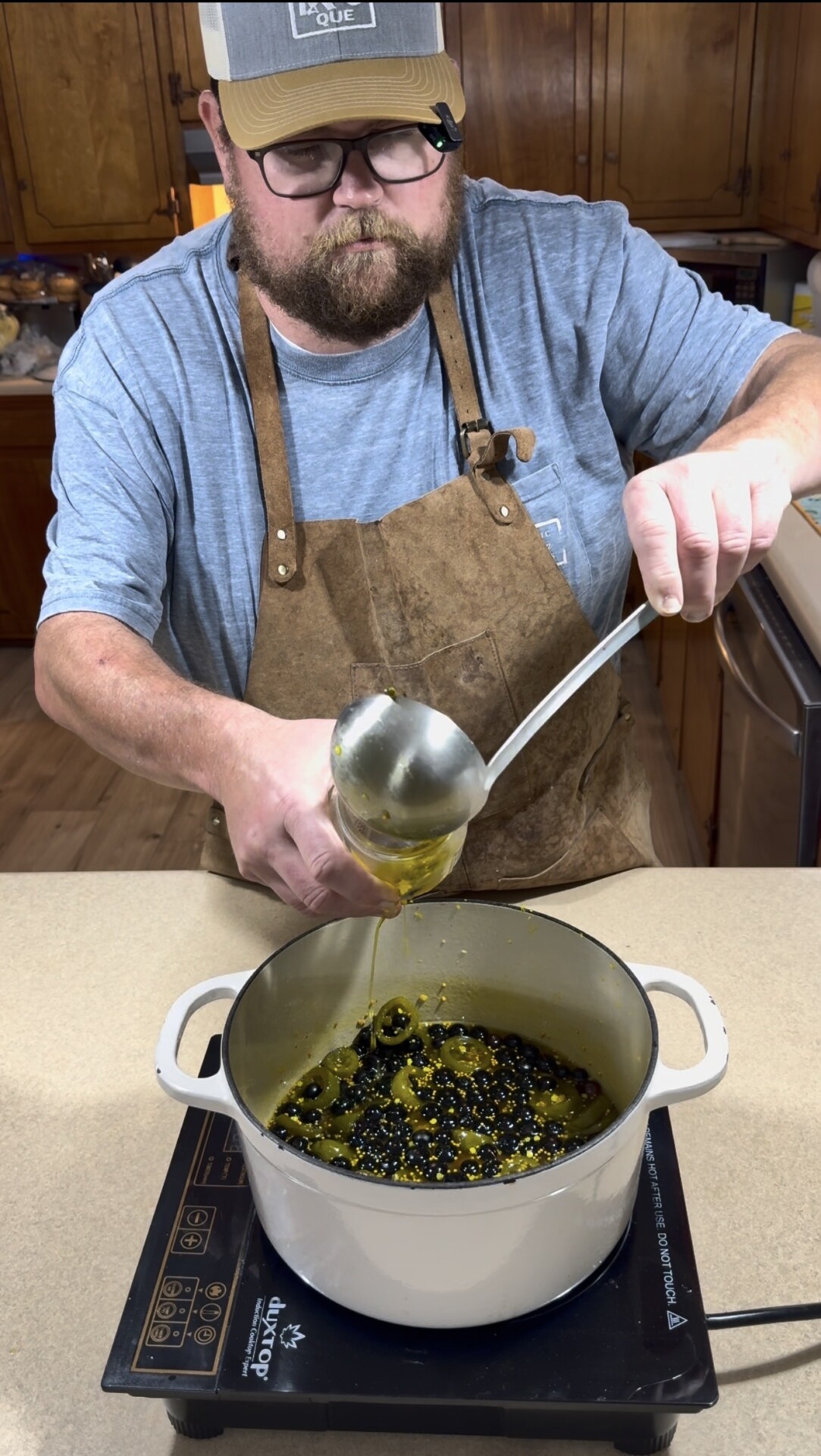 A bearded man wearing glasses, a brown apron, and a blue shirt pours a yellow liquid from a ladle into a white pot filled with beans on a stovetop in a kitchen.