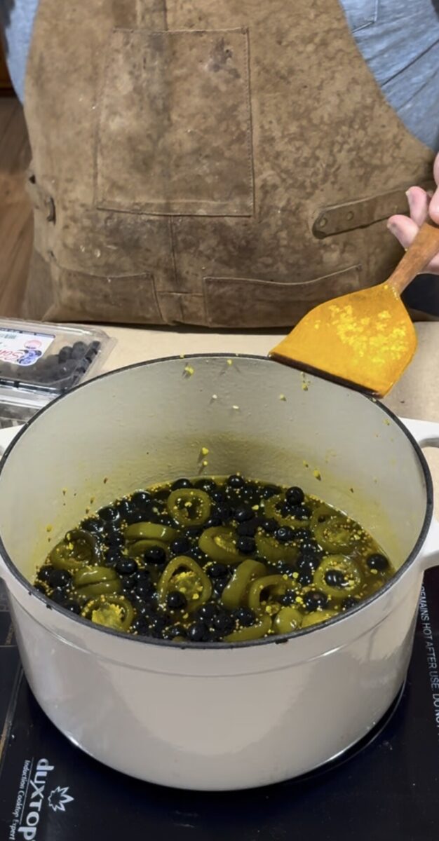 A person in a brown apron stirs a pot filled with sliced jalapeños, blueberries, and liquid on a stovetop. A container of fresh blueberries is visible beside the pot.