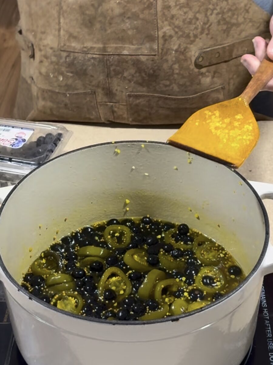 A person wearing a brown apron stirs a white pot filled with sliced jalapeños and blueberries using a yellow spatula. A container of blueberries sits nearby on the counter.