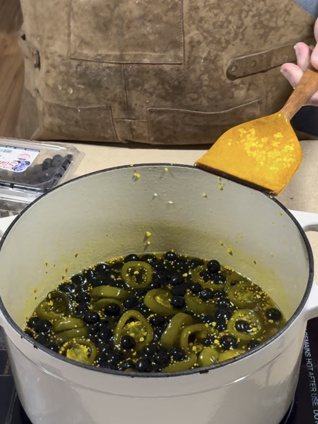 A person wearing a brown apron stirs a white pot filled with sliced jalapeños and blueberries using a yellow spatula. A container of blueberries sits nearby on the counter.