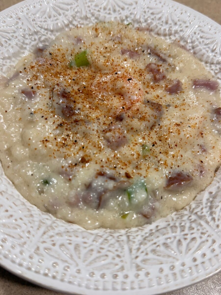 A bowl of creamy grits mixed with pieces of corned beef and green onions, topped with a generous sprinkle of seasoning, served in a decorative white bowl.