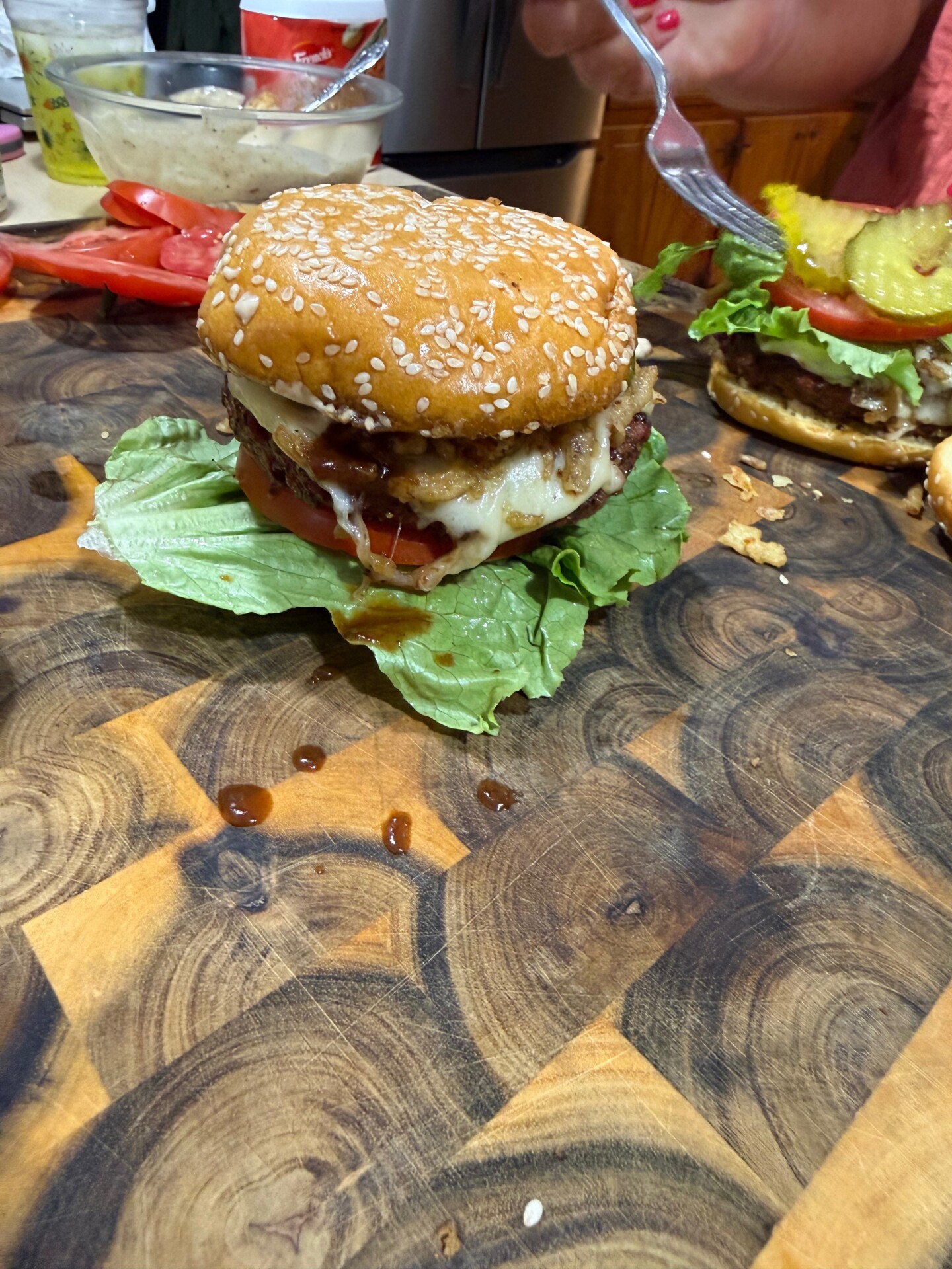 A cheeseburger with lettuce, tomato, and pickles sits on a wooden cutting board. Nearby, a person with red nail polish holds a fork over another burger in the background.