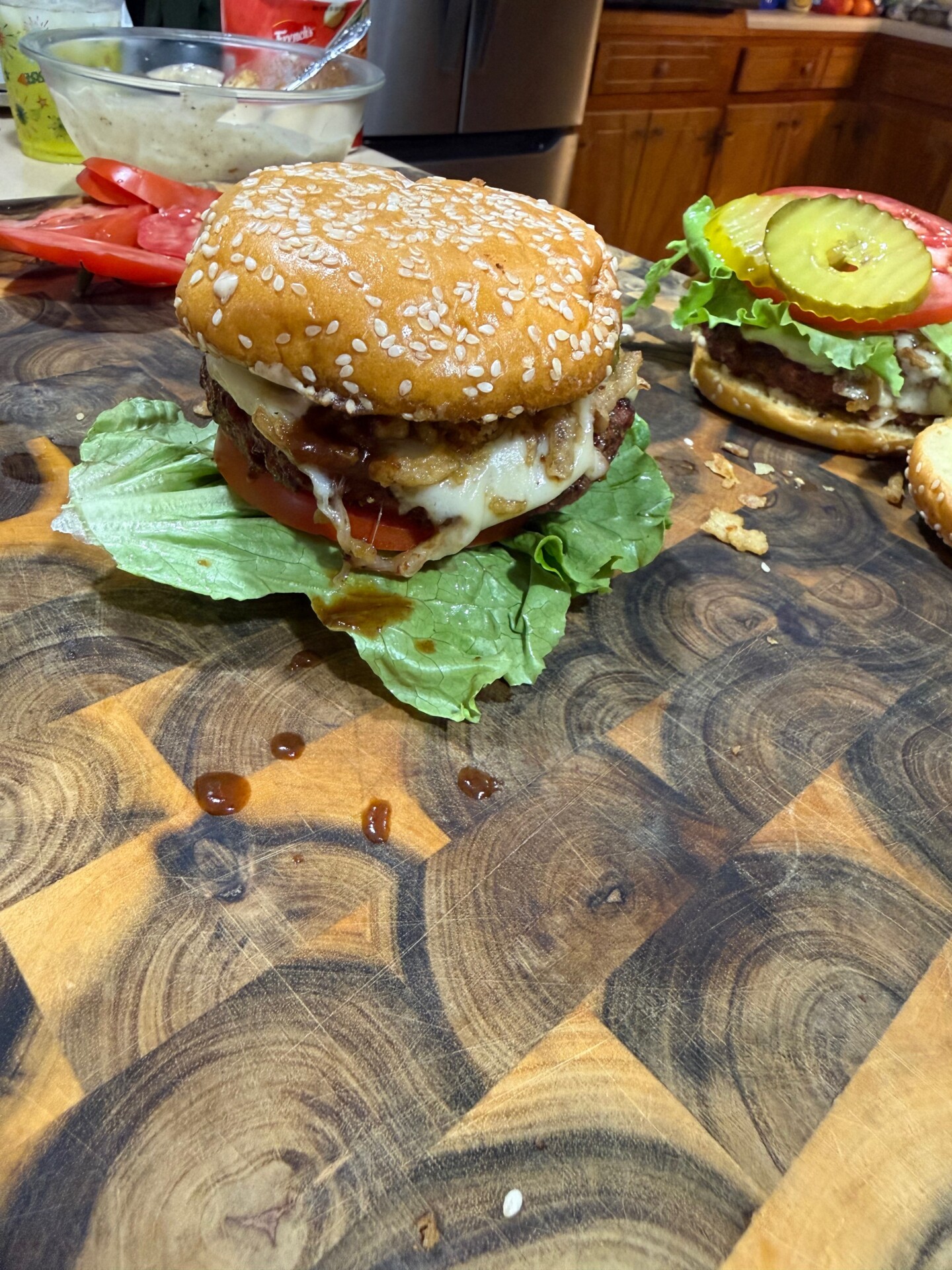 A close-up of a homemade cheeseburger with lettuce, tomato, melted cheese, and sauce on a sesame seed bun, resting on a wooden cutting board. Another burger and sliced tomatoes are visible in the background.