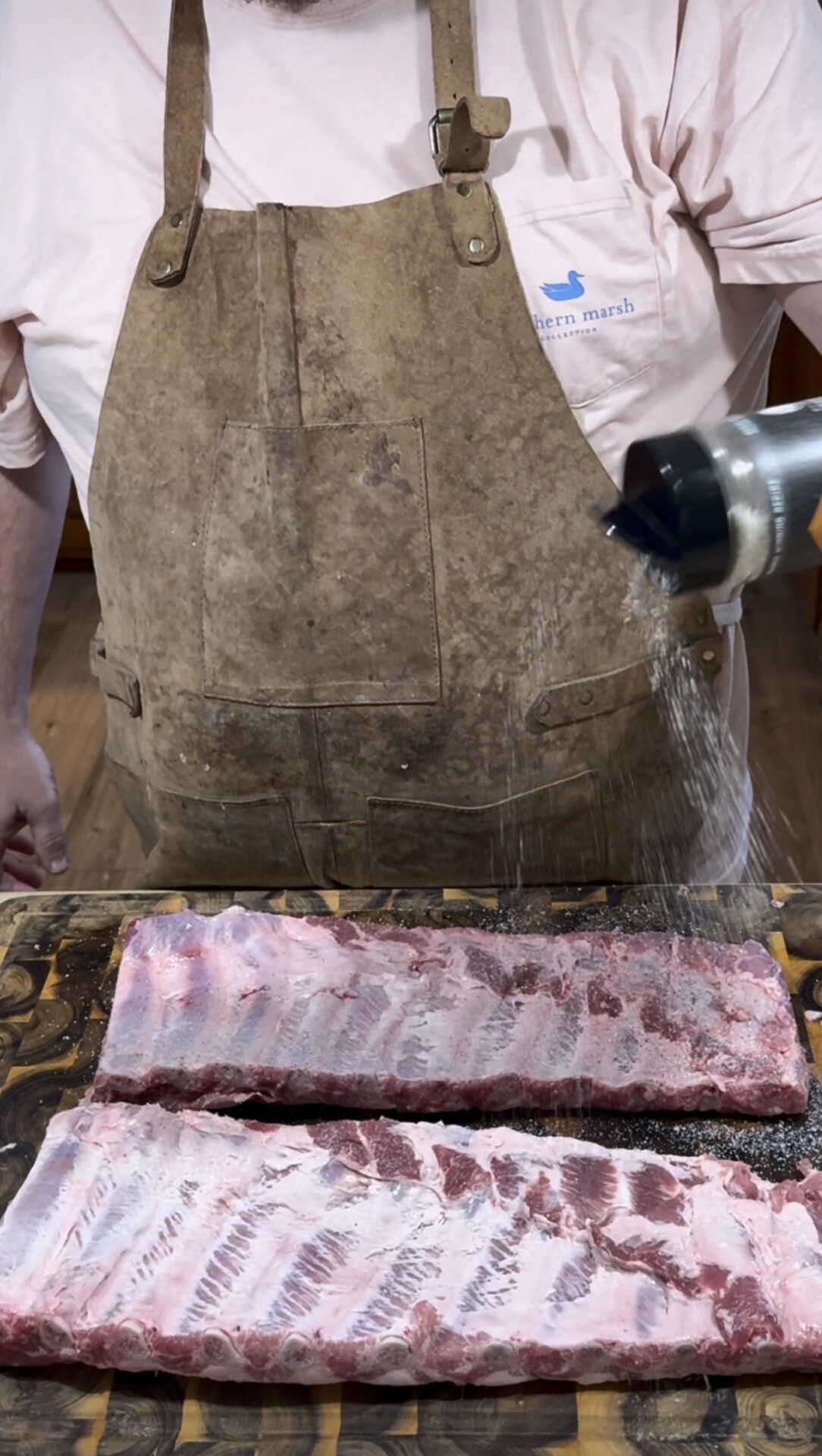 A person wearing a brown apron and pink shirt sprinkles seasoning from a shaker onto two raw racks of ribs placed on a wooden cutting board.