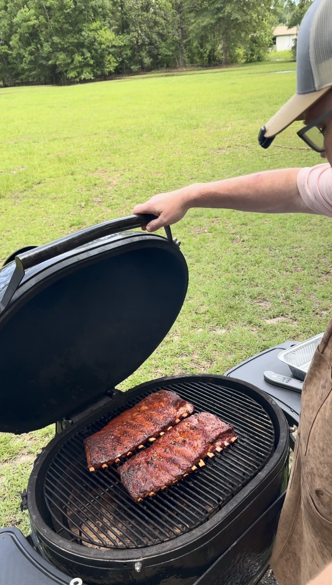 A person wearing a cap and apron lifts the lid of a grill, revealing two racks of seasoned ribs cooking on the grate. The scene is outdoors, with green grass and trees in the background.