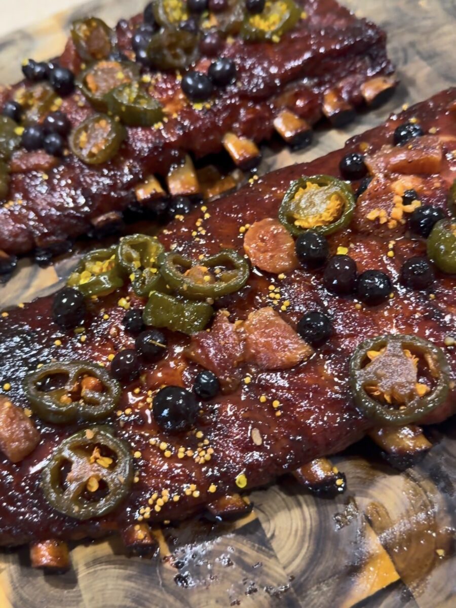Close-up of two racks of glazed ribs topped with sliced jalapeños, blueberries, and bits of bacon, displayed on a wooden cutting board. The ribs appear juicy with a shiny, caramelized glaze.