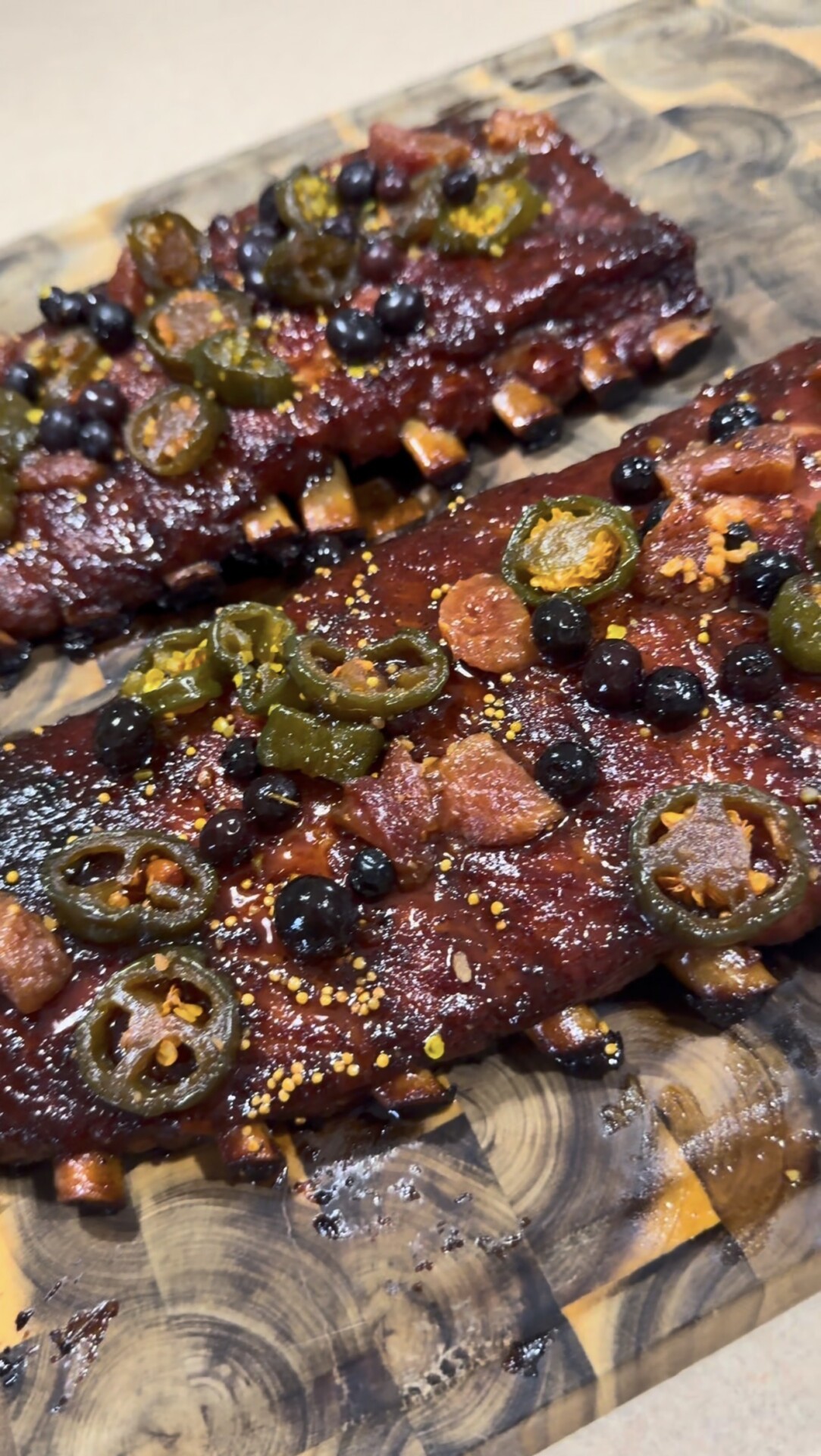 Two racks of glazed ribs topped with sliced jalapeños, blueberries, and small pieces of bacon, served on a wooden cutting board.