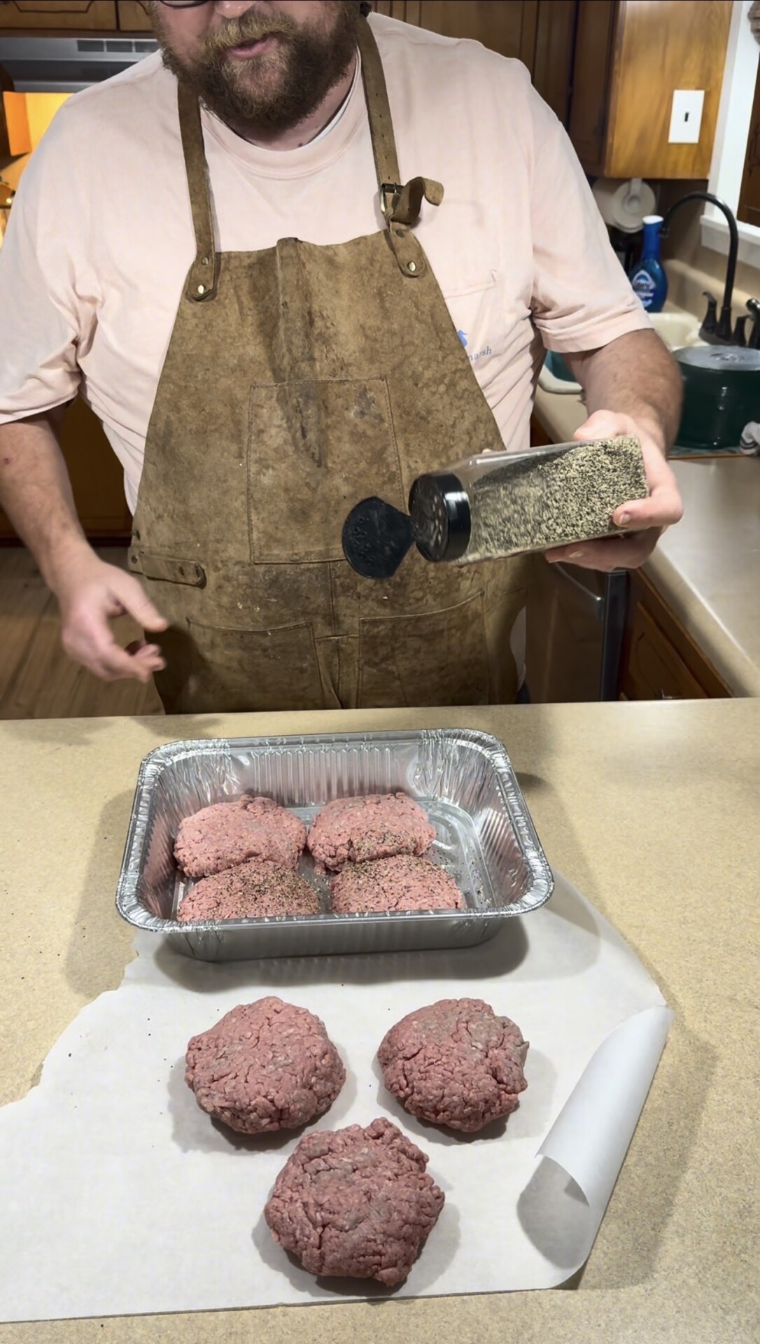A person in a brown apron seasons raw burger patties with spices. Four patties are in a metal tray, and three more are on parchment paper on a countertop in a kitchen setting.