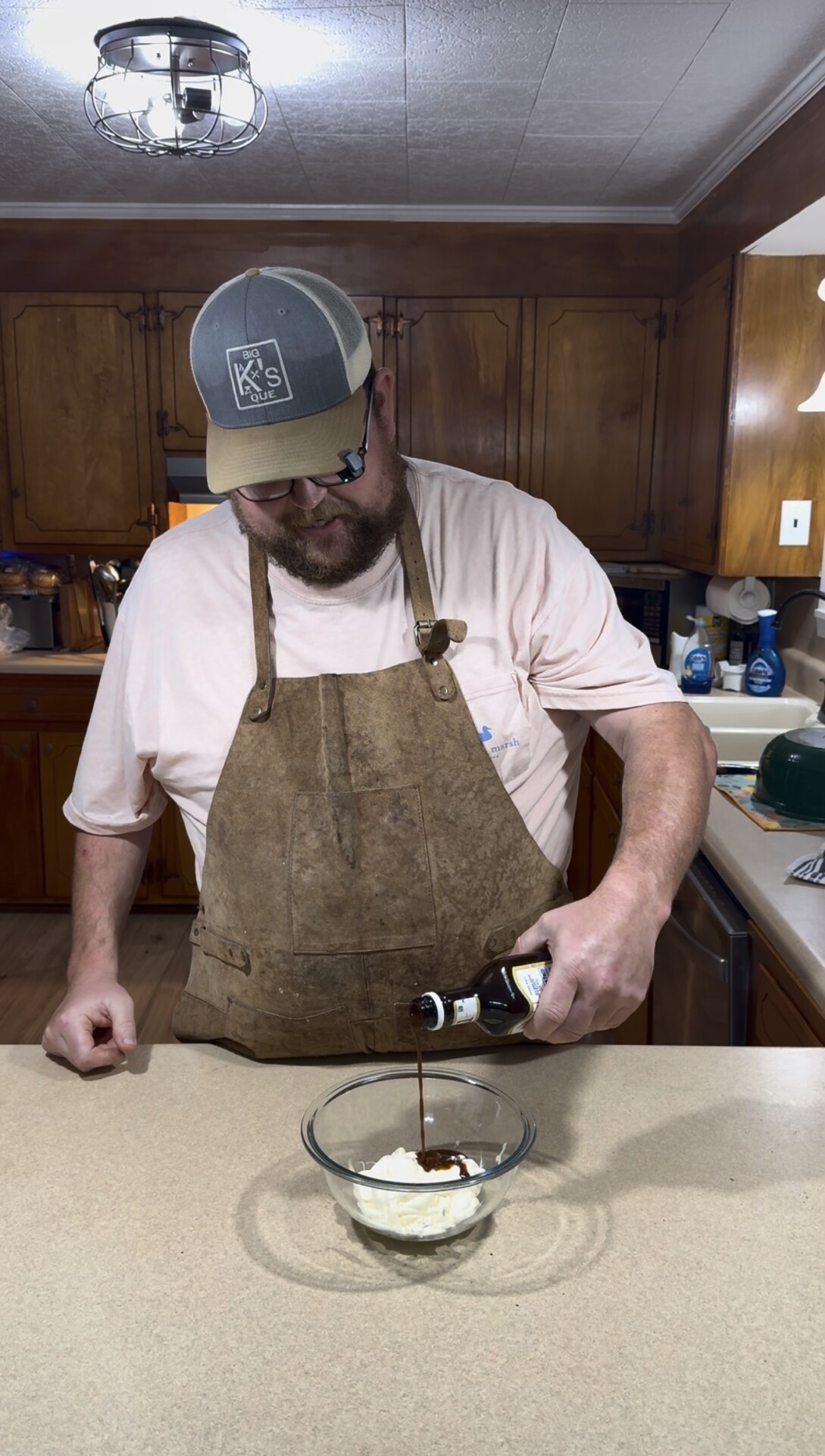 A man wearing a brown apron and gray cap pours liquid from a bottle into a glass bowl with a white substance on a kitchen counter. Wooden cabinets and kitchen items are visible in the background.