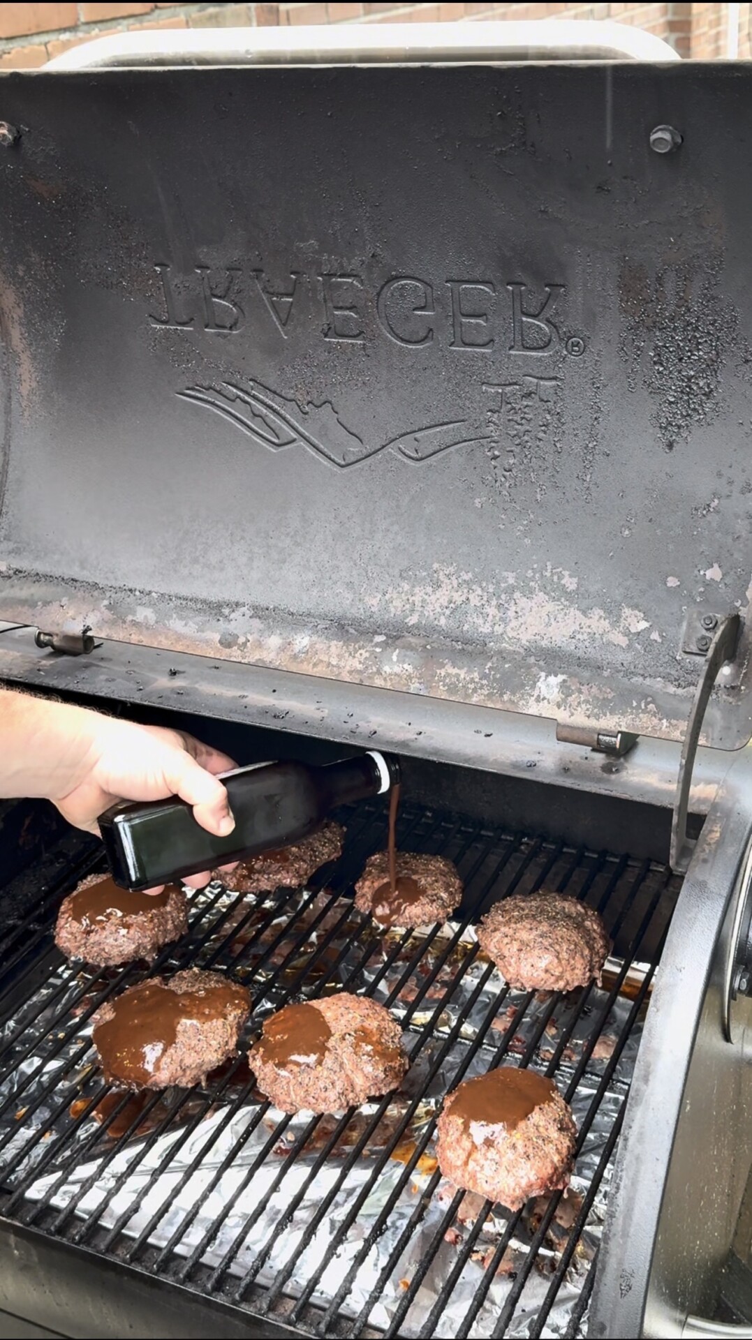 A hand pours barbecue sauce from a dark bottle onto several hamburger patties cooking on the grates of a Traeger smoker grill. Steam rises from the burgers as they cook.