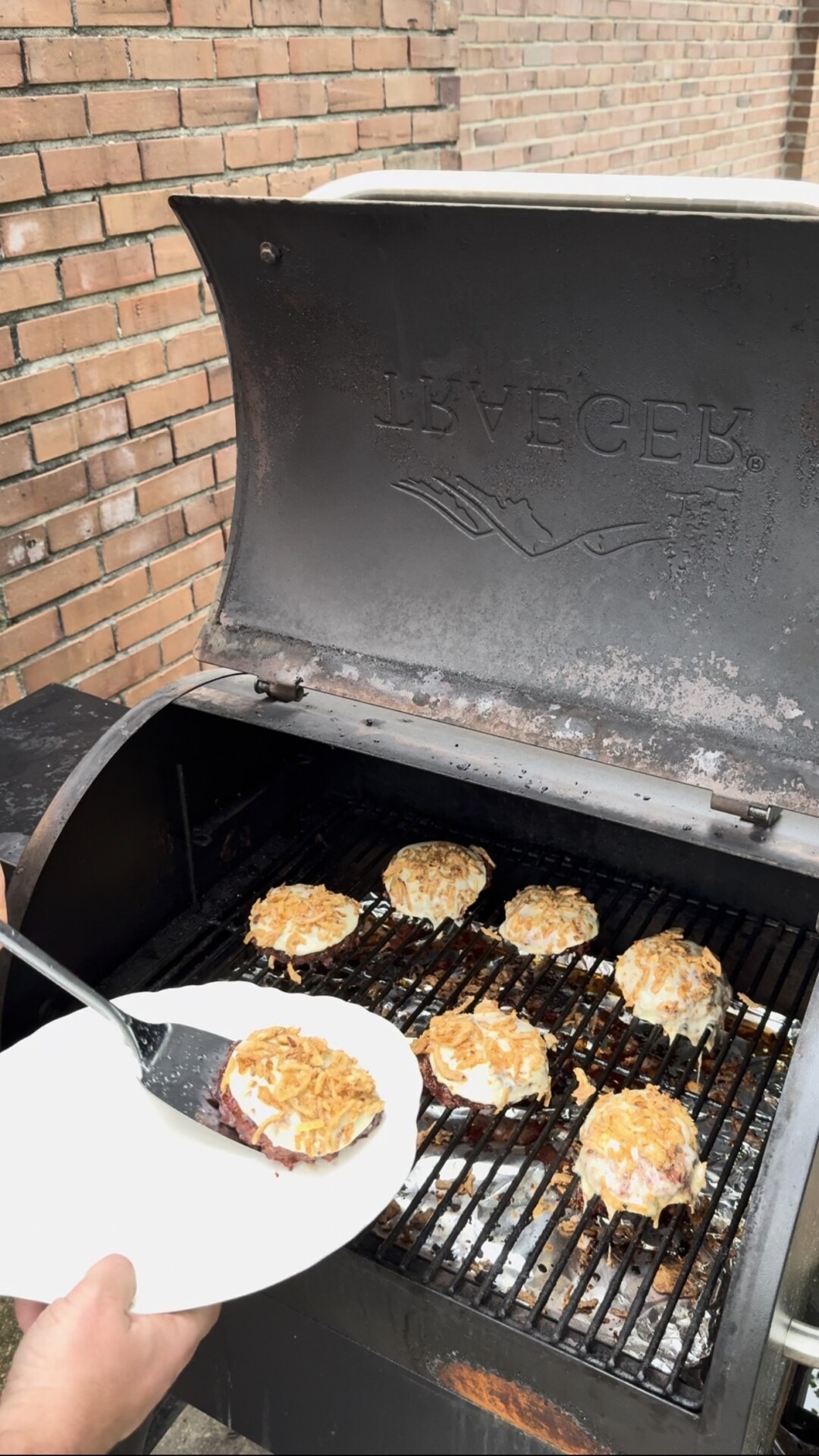 A person removes a grilled burger patty topped with melted cheese and crispy onions from a smoker grill onto a plate. Other similar patties are still cooking on the grill, next to a brick wall.